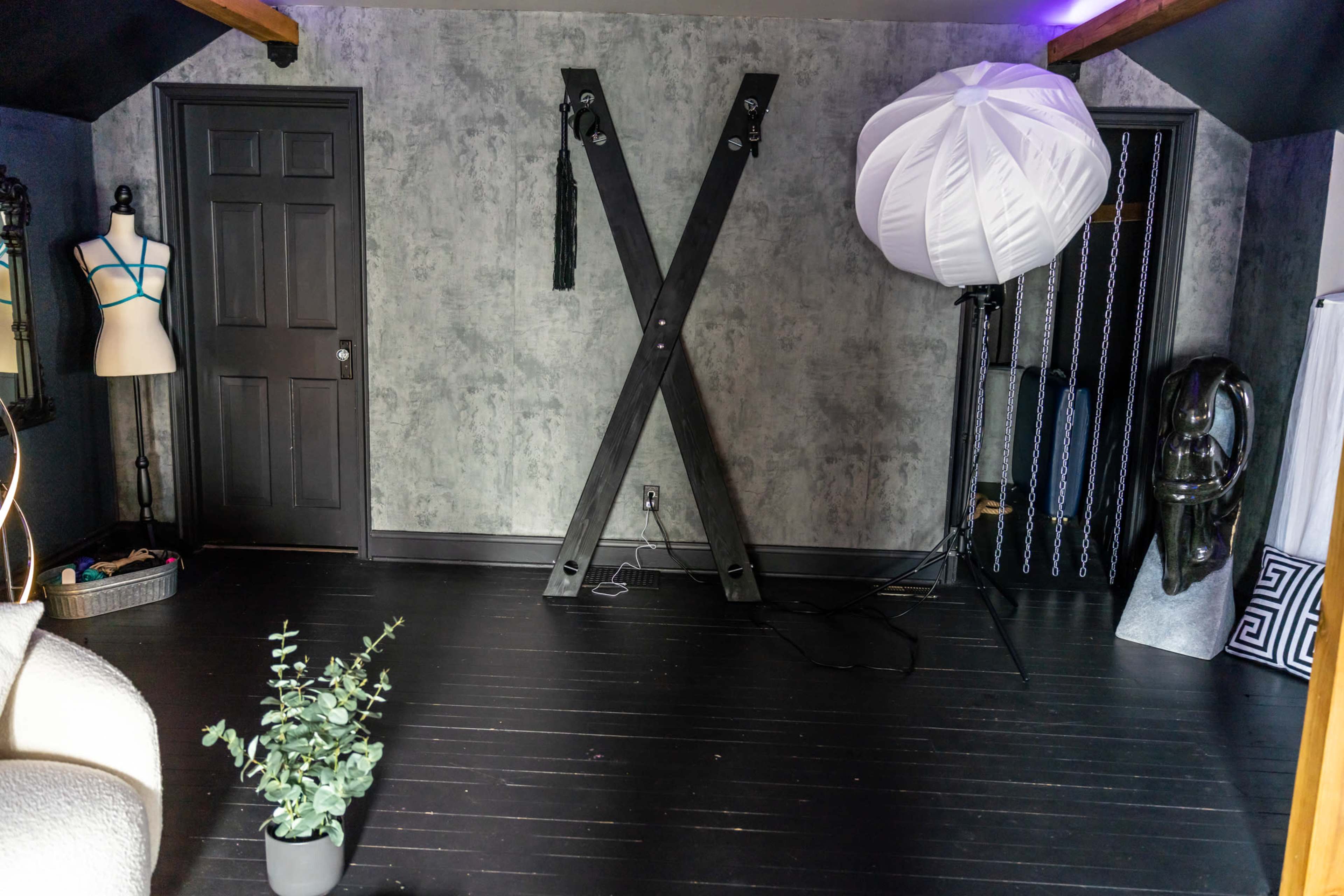 An indoor studio space featuring a large black X-frame, a photography light umbrella, a mannequin, and various accessories against a textured gray wall.