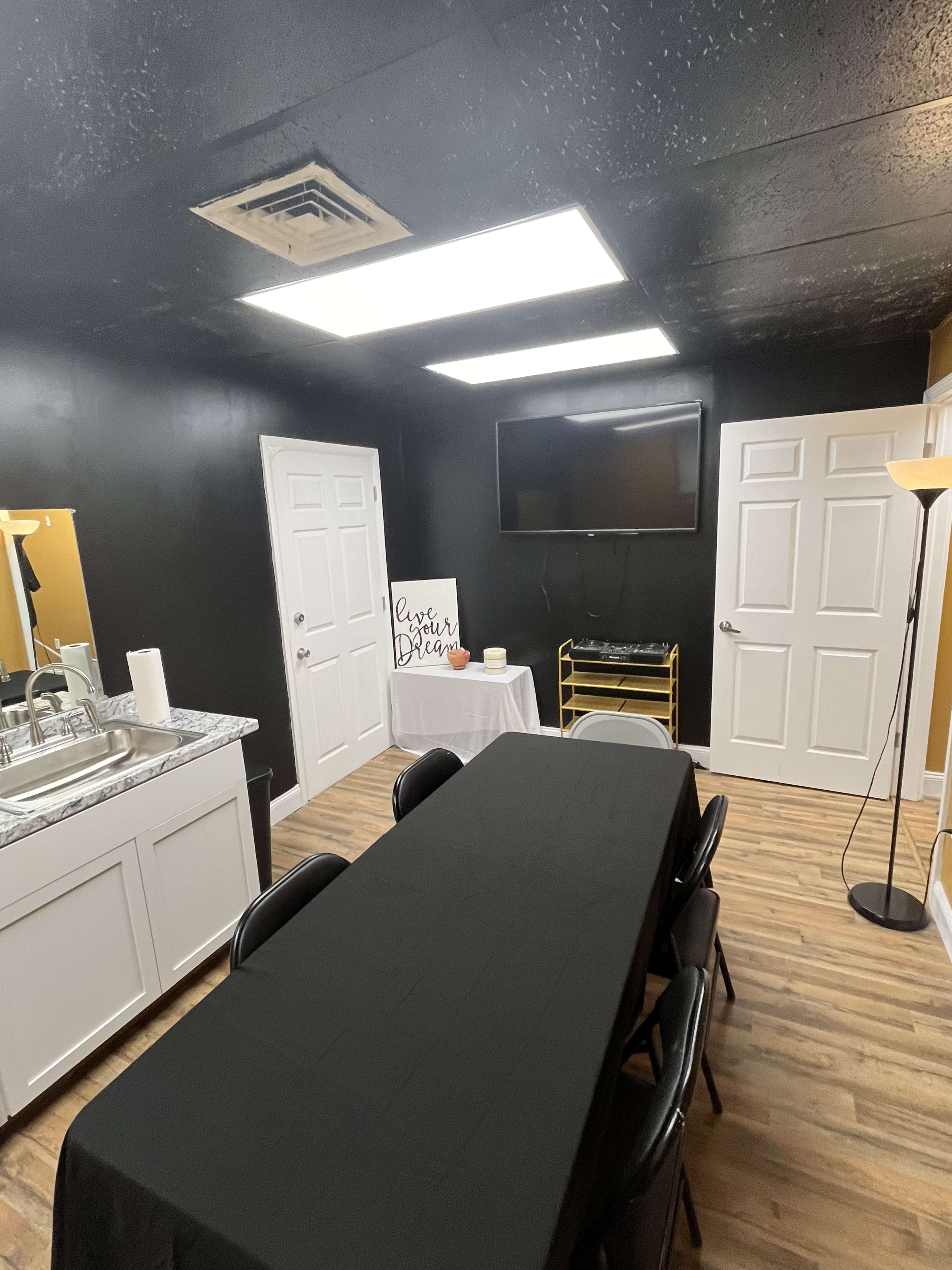 A small room features a long black table, a television on the wall, and a sink area, with black walls and wooden flooring.