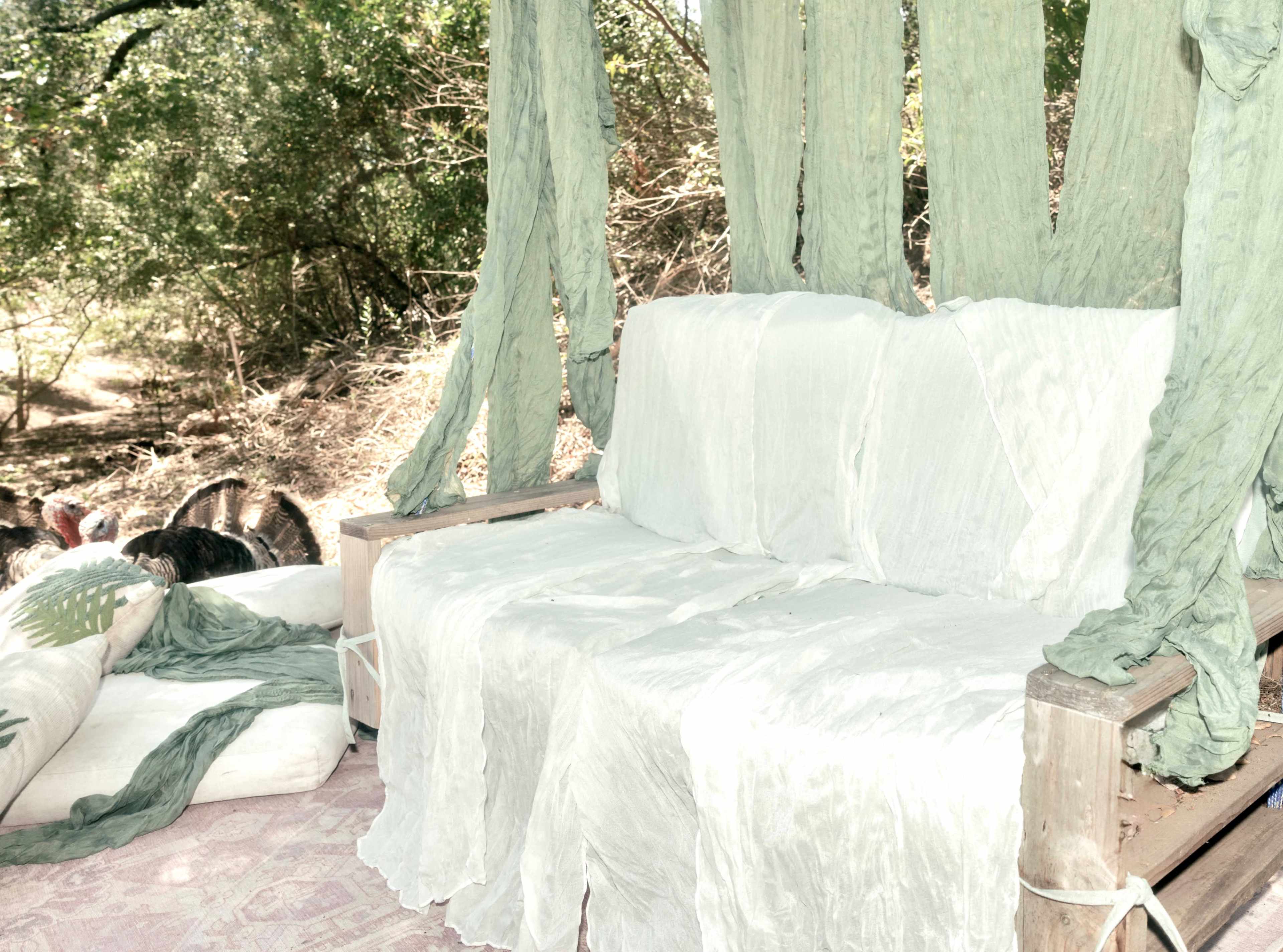 A wooden bench is covered with white and green fabric, surrounded by a natural setting with trees and greenery.