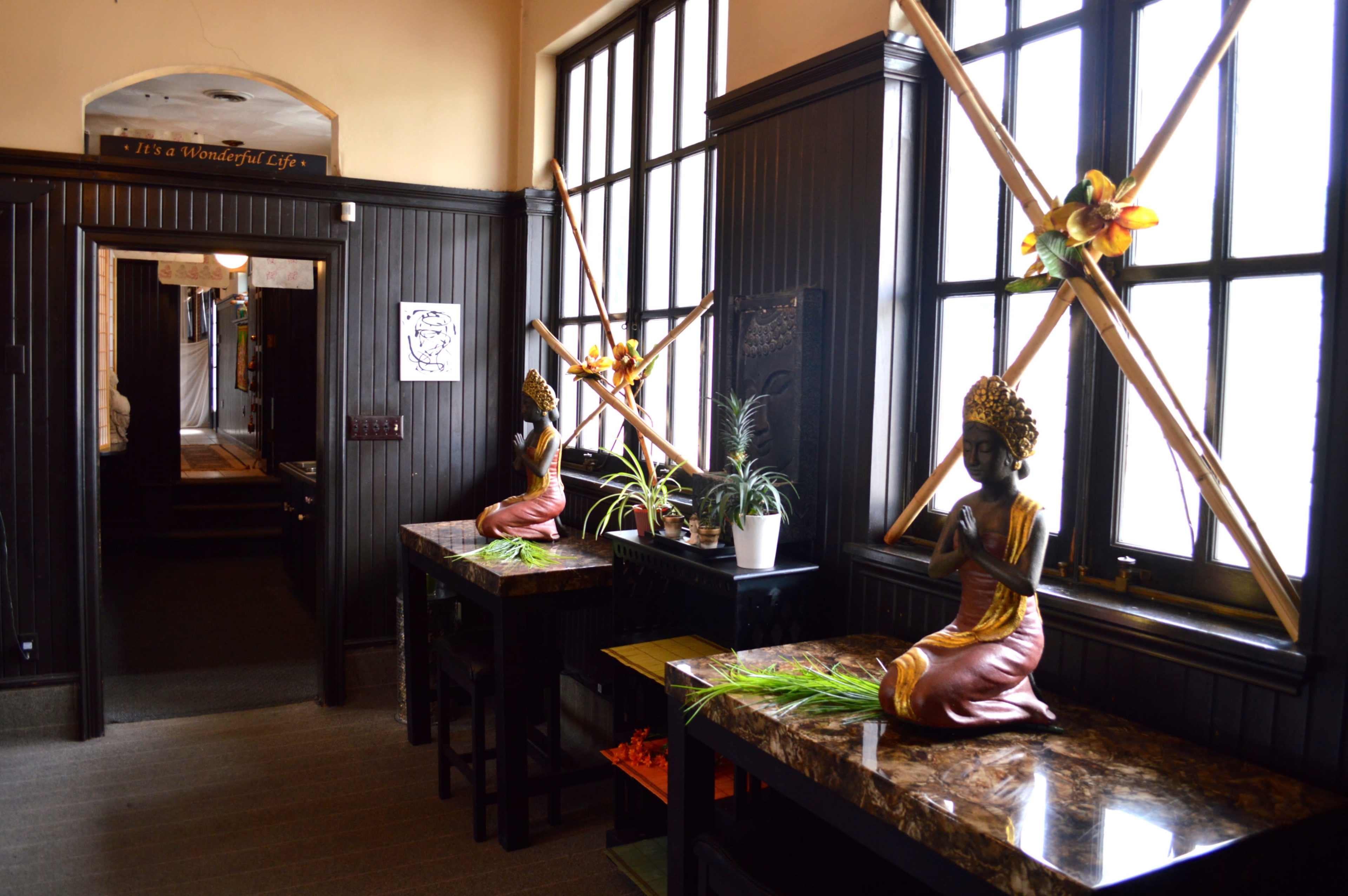 A hallway with two decorative statues and plants on black tables, framed by large windows with wooden cross beams.