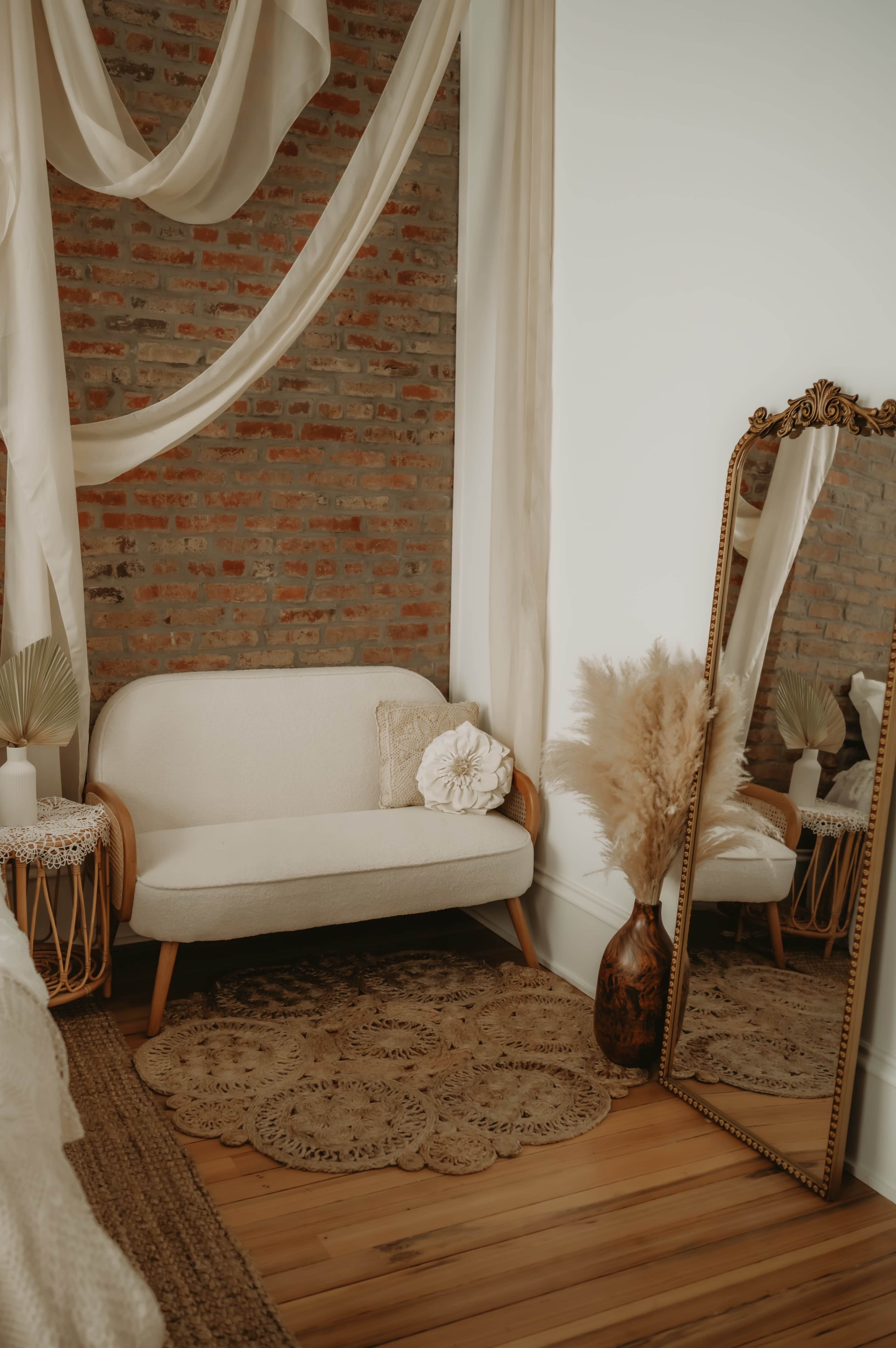 The image shows a cozy corner of a room featuring a light-colored loveseat, a tall mirror with an ornate frame, and decorative elements like a vase of dried flowers and a textured rug against a brick wall.