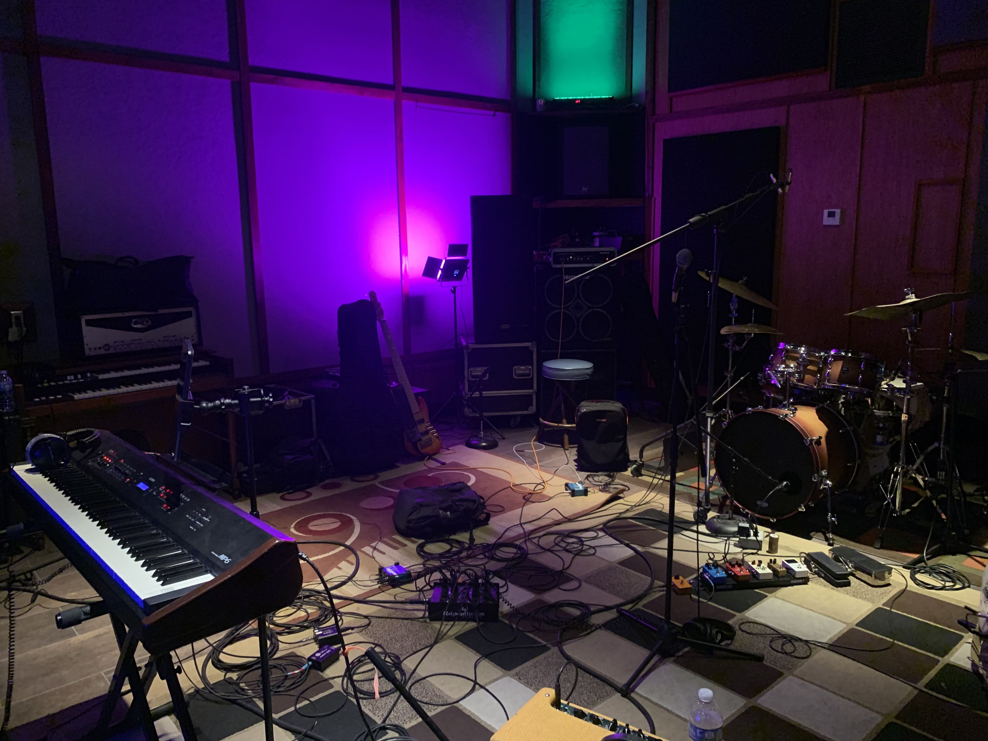 A music studio with a keyboard, microphone stand, guitar, amplifiers, and a drum set surrounded by cables and equipment.