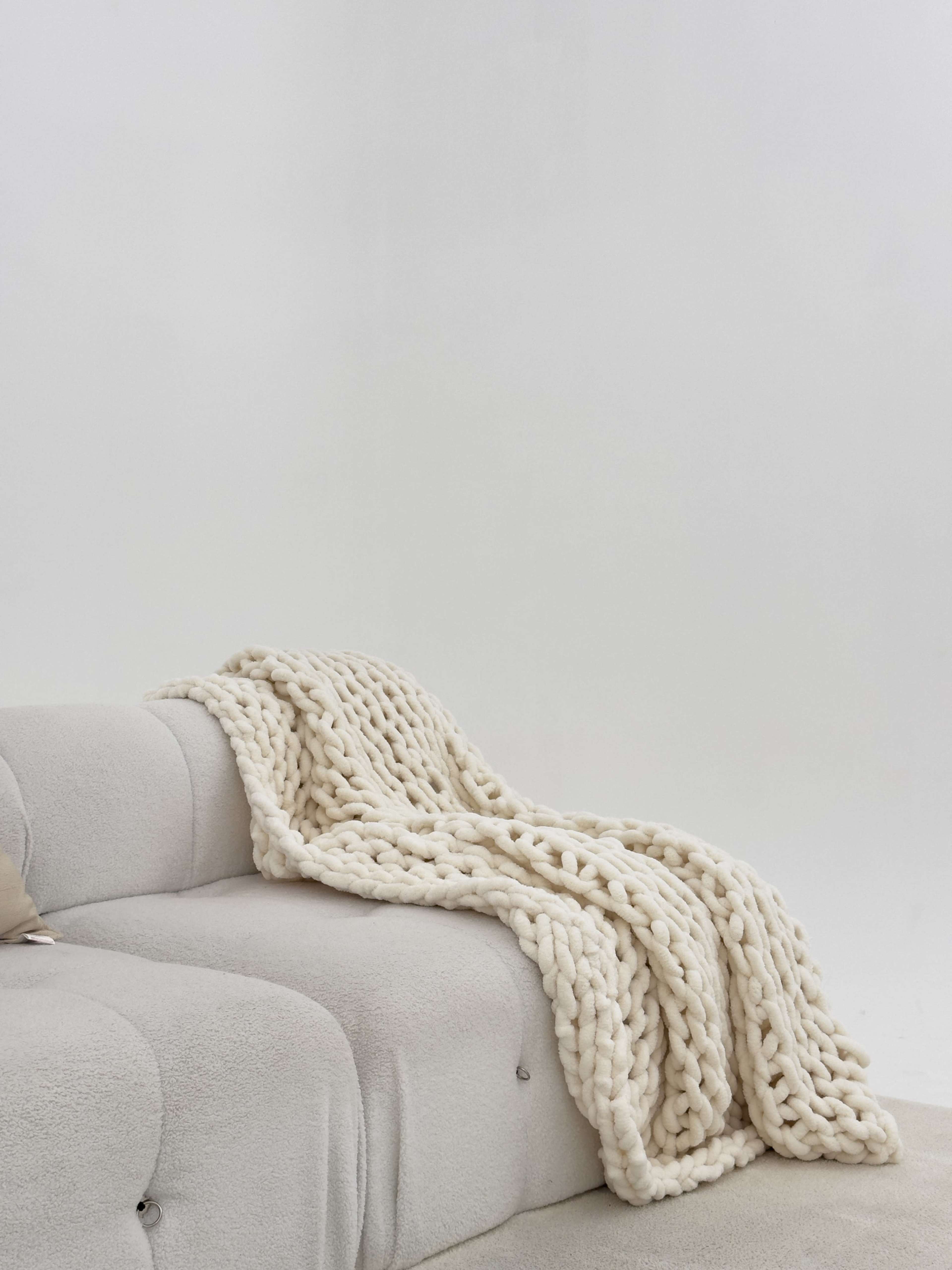 A cream-colored chunky knit blanket is draped over a light grey couch in a minimalistic setting.