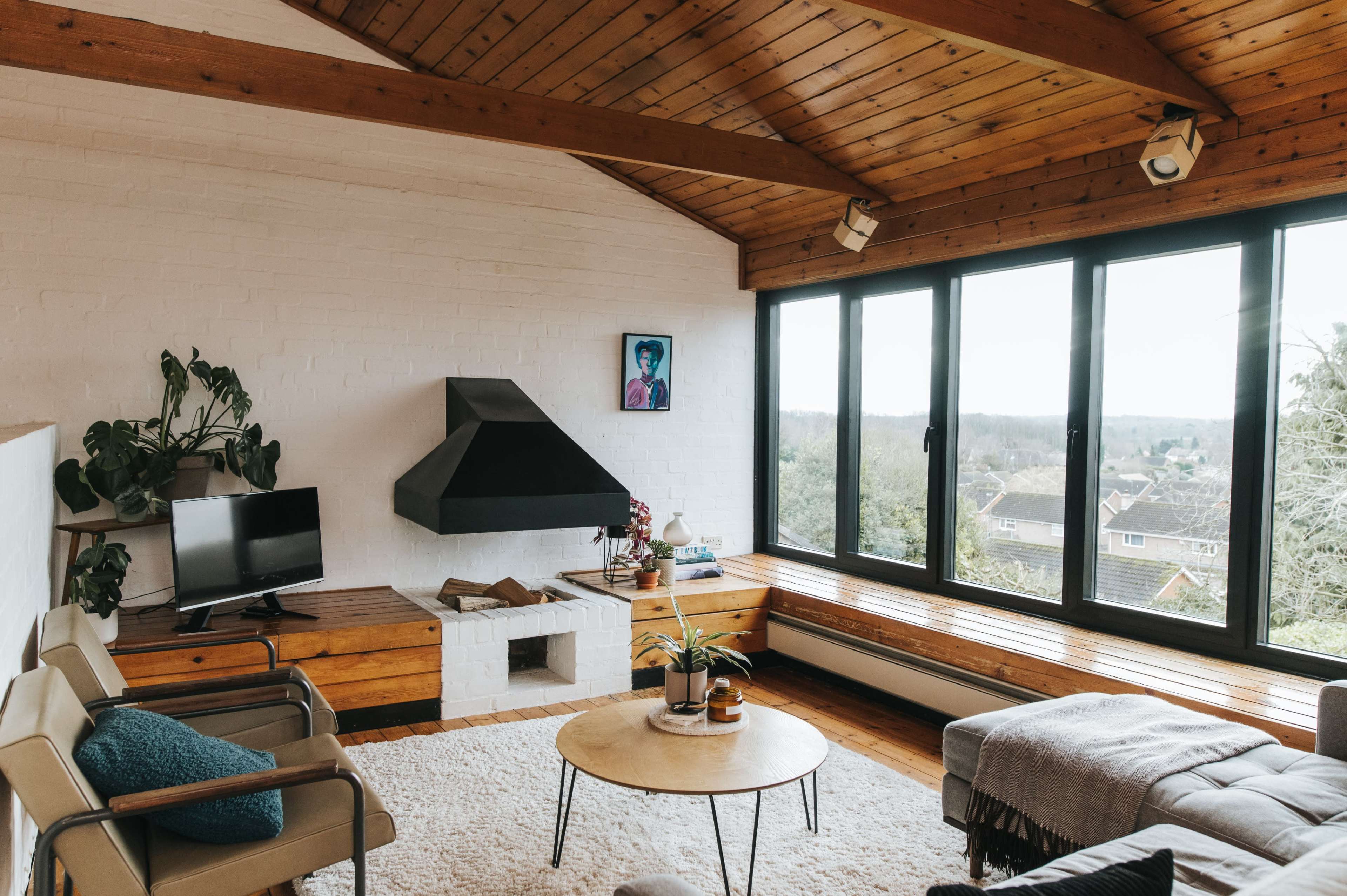 A modern living room features a wooden ceiling, large windows overlooking a hillside, a fireplace, and minimalistic furniture.