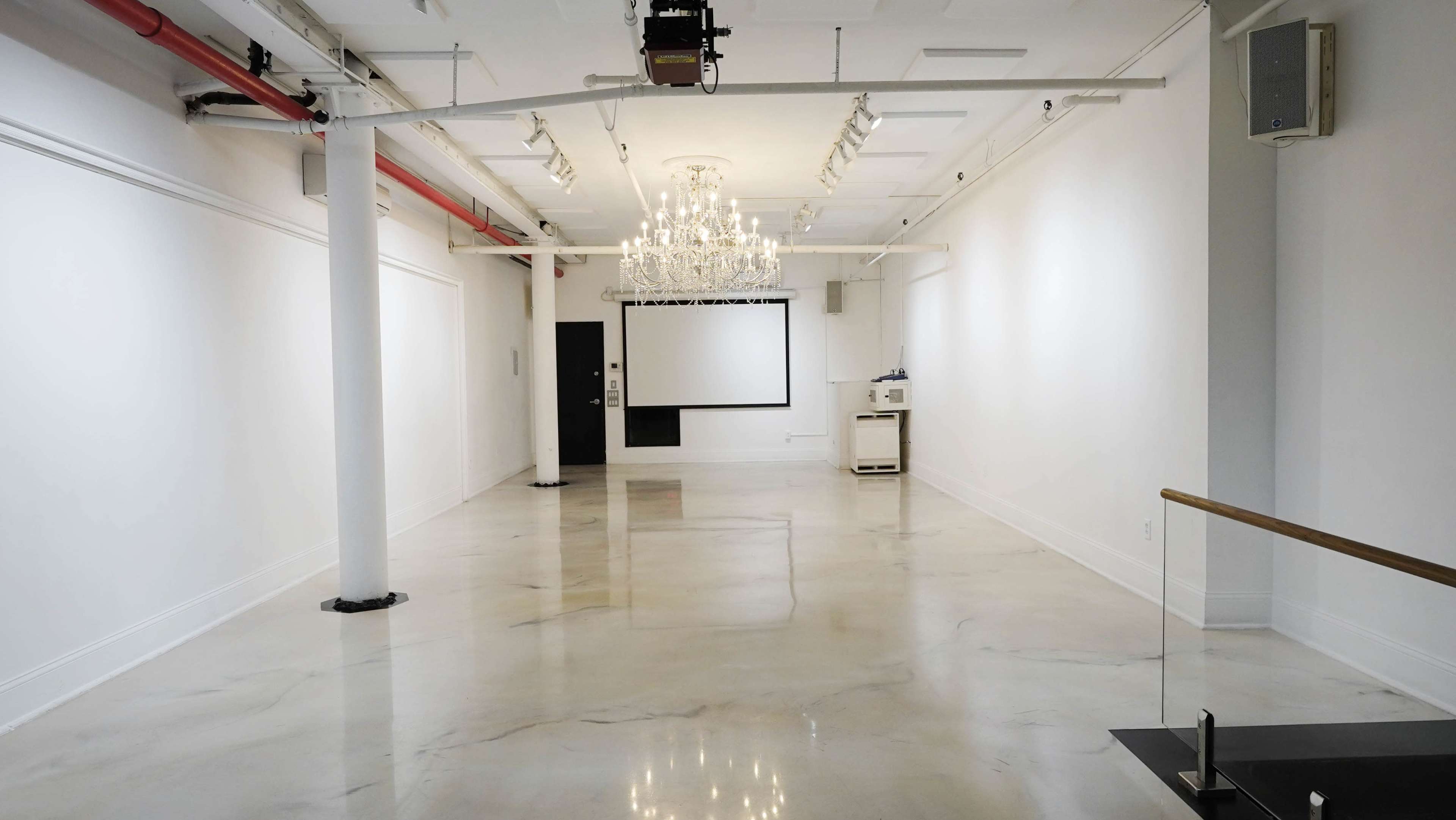The image shows a spacious, empty gallery with polished concrete floors and white walls, featuring a chandelier and a projection screen at the far end.