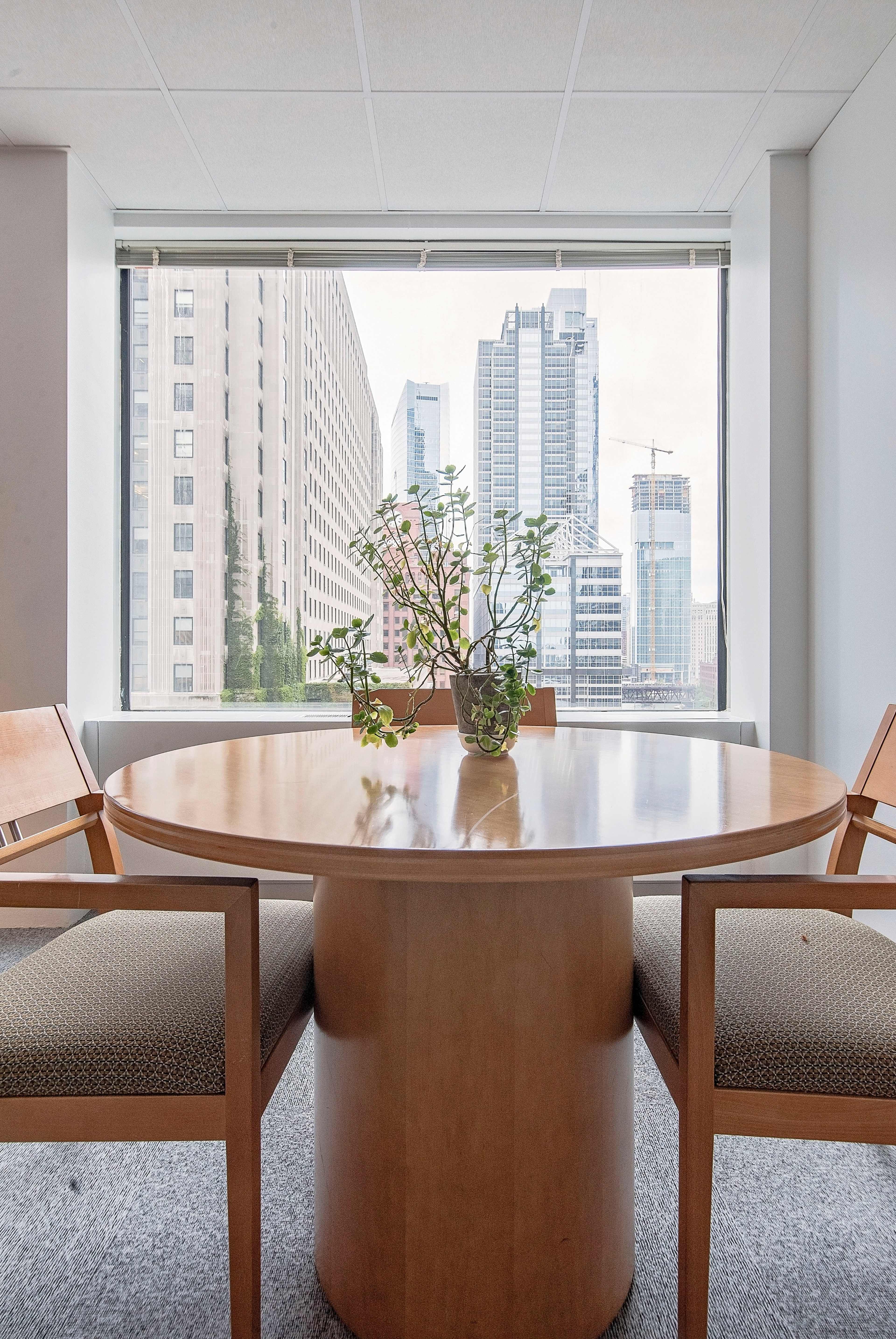 A round wooden table with two chairs sits in an office space, featuring a view of tall city buildings through a large window.