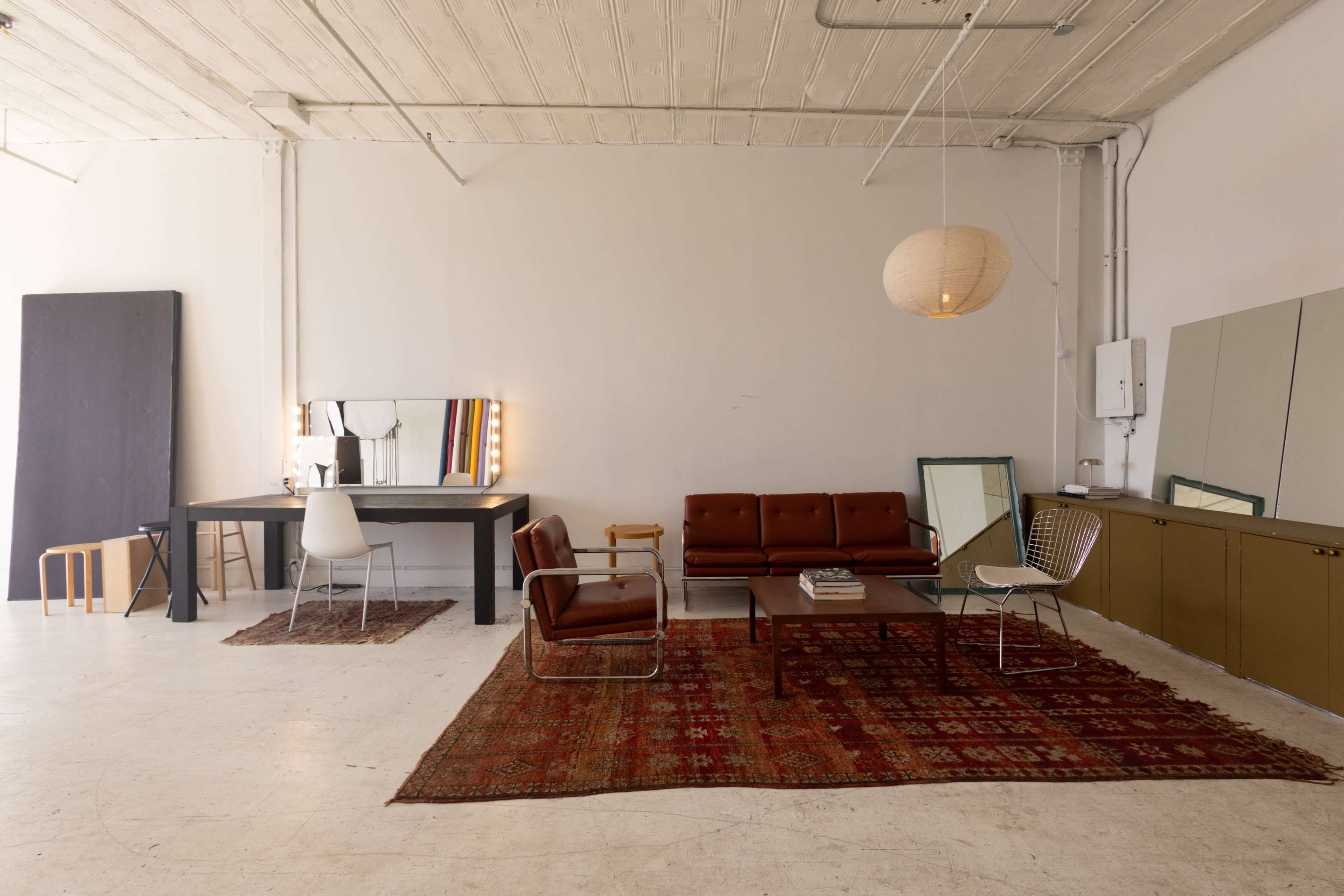 The image shows a spacious room with a mix of modern and vintage furniture, including a brown leather sofa, a large table, and various mirrors arranged on the walls.