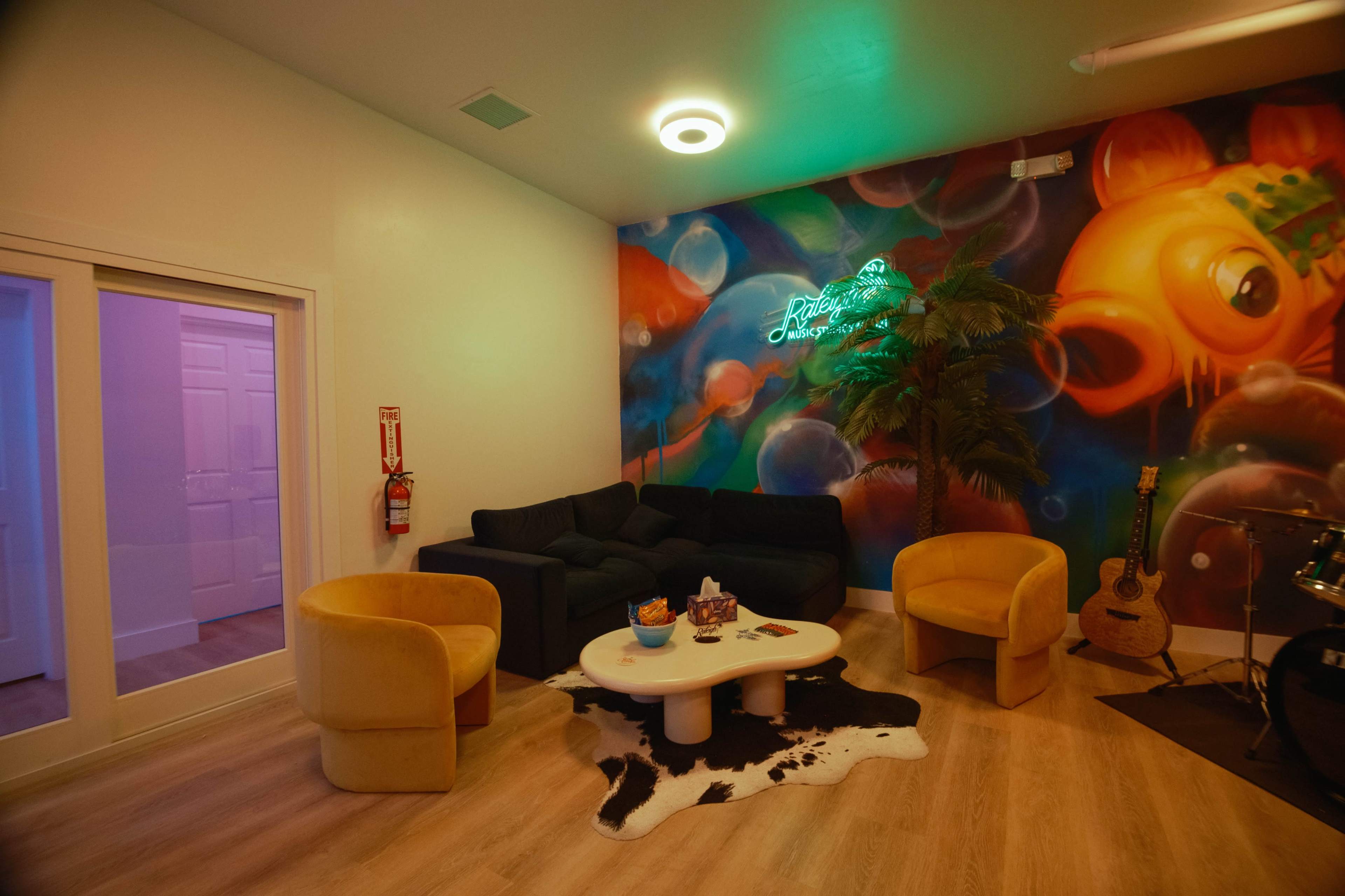 The image depicts a vibrant lounge area with colorful wall art, a dark sofa, yellow seating, a coffee table, and a decorative rug.