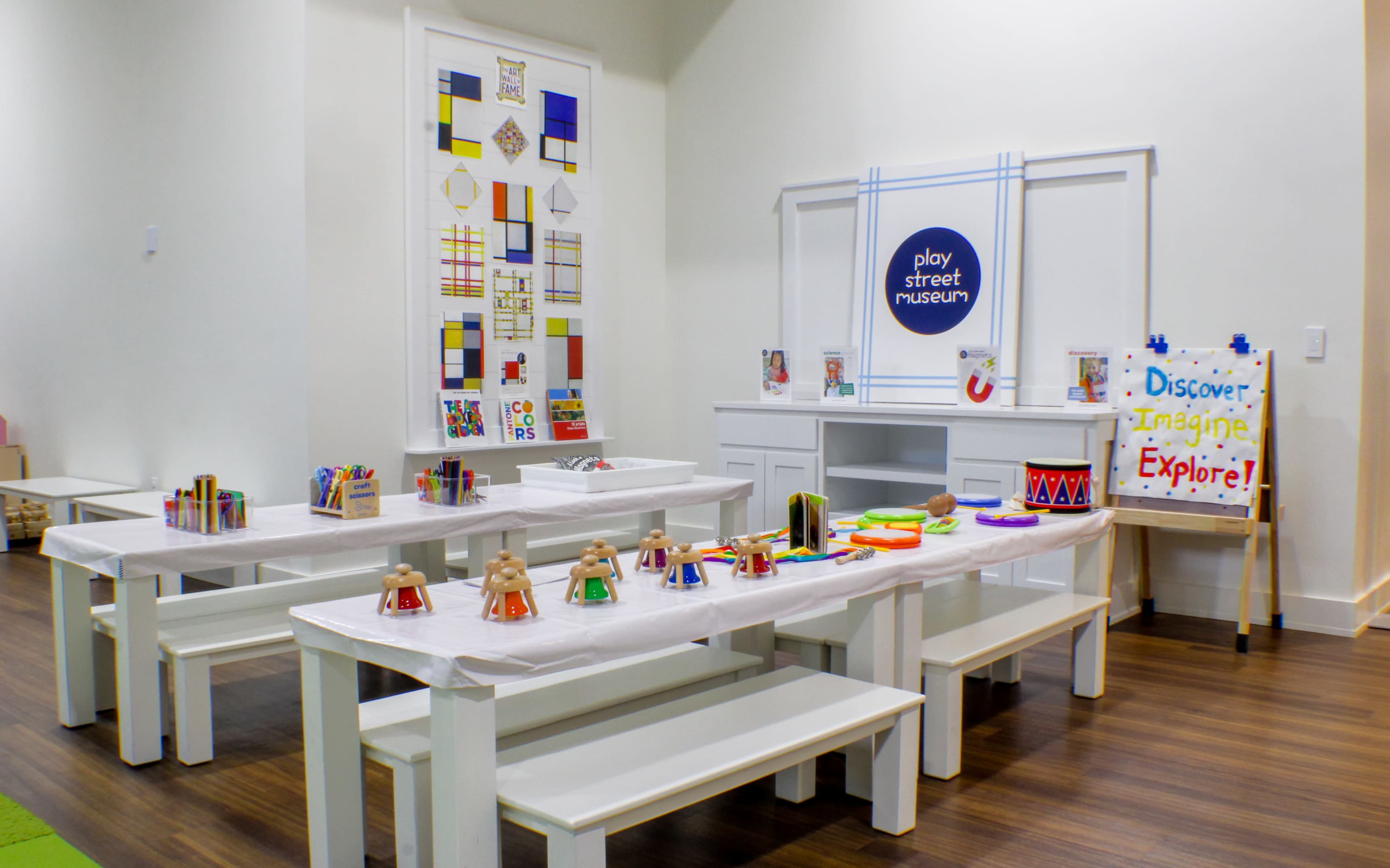 A colorful children's art and activity room with white tables, benches, and various craft supplies arranged on the tables.