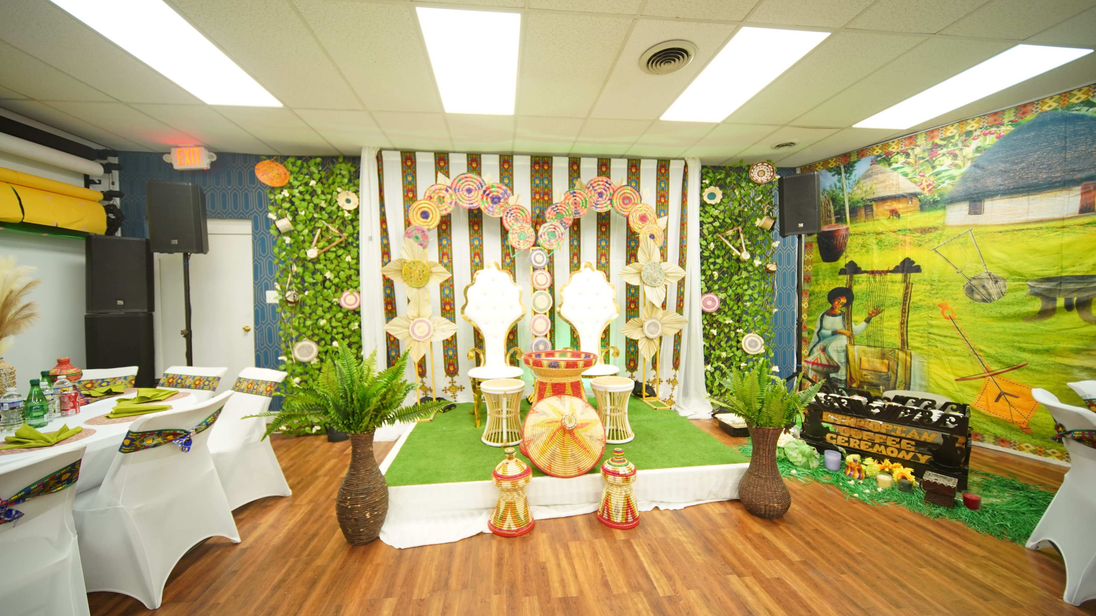 The image shows a decorated indoor space with colorful circular decorations, greenery on the walls, and two chairs set on a turf-like surface, flanked by potted plants.