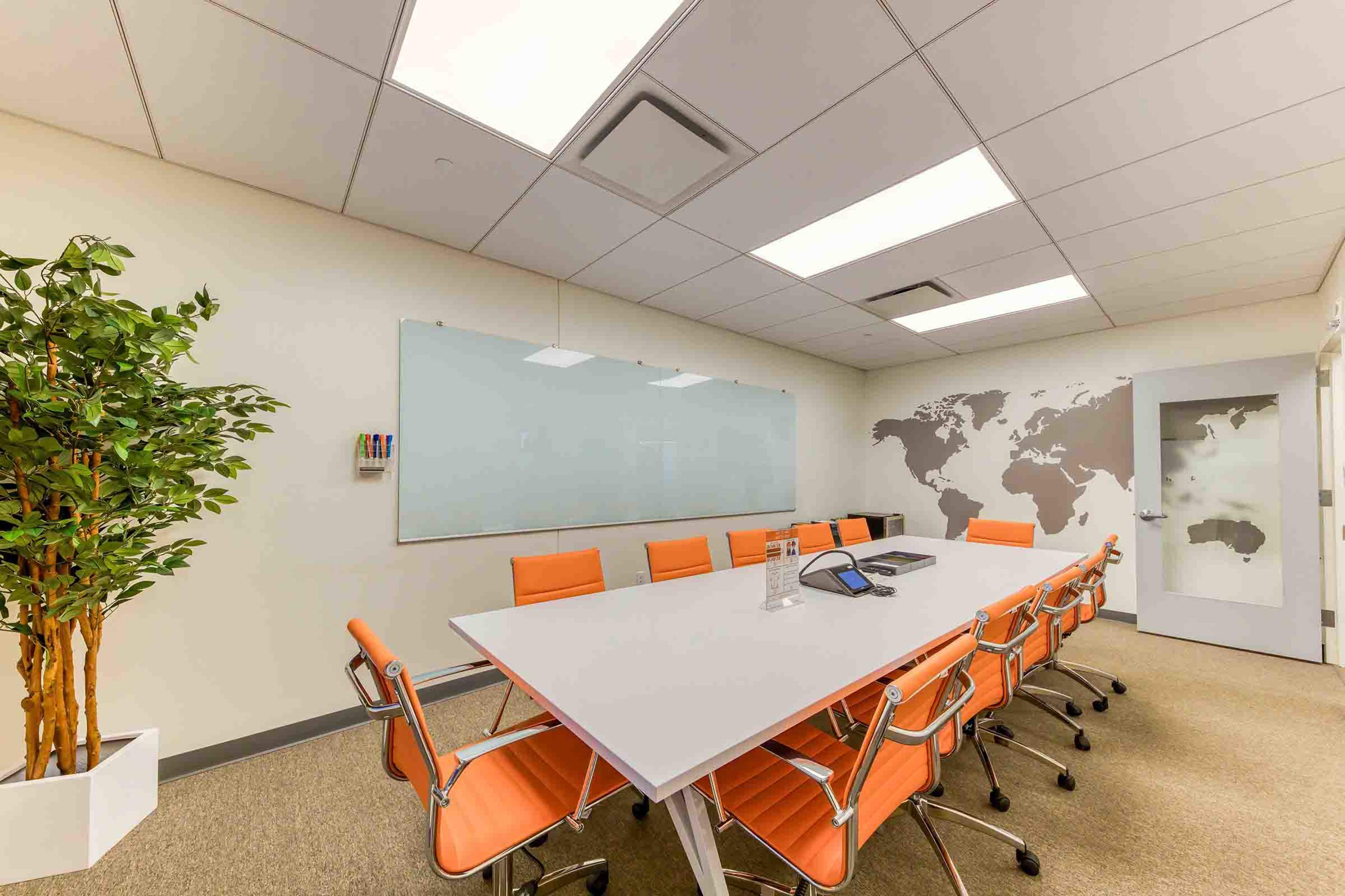 A modern conference room features a long white table surrounded by orange chairs, with a large whiteboard and a world map on the wall.