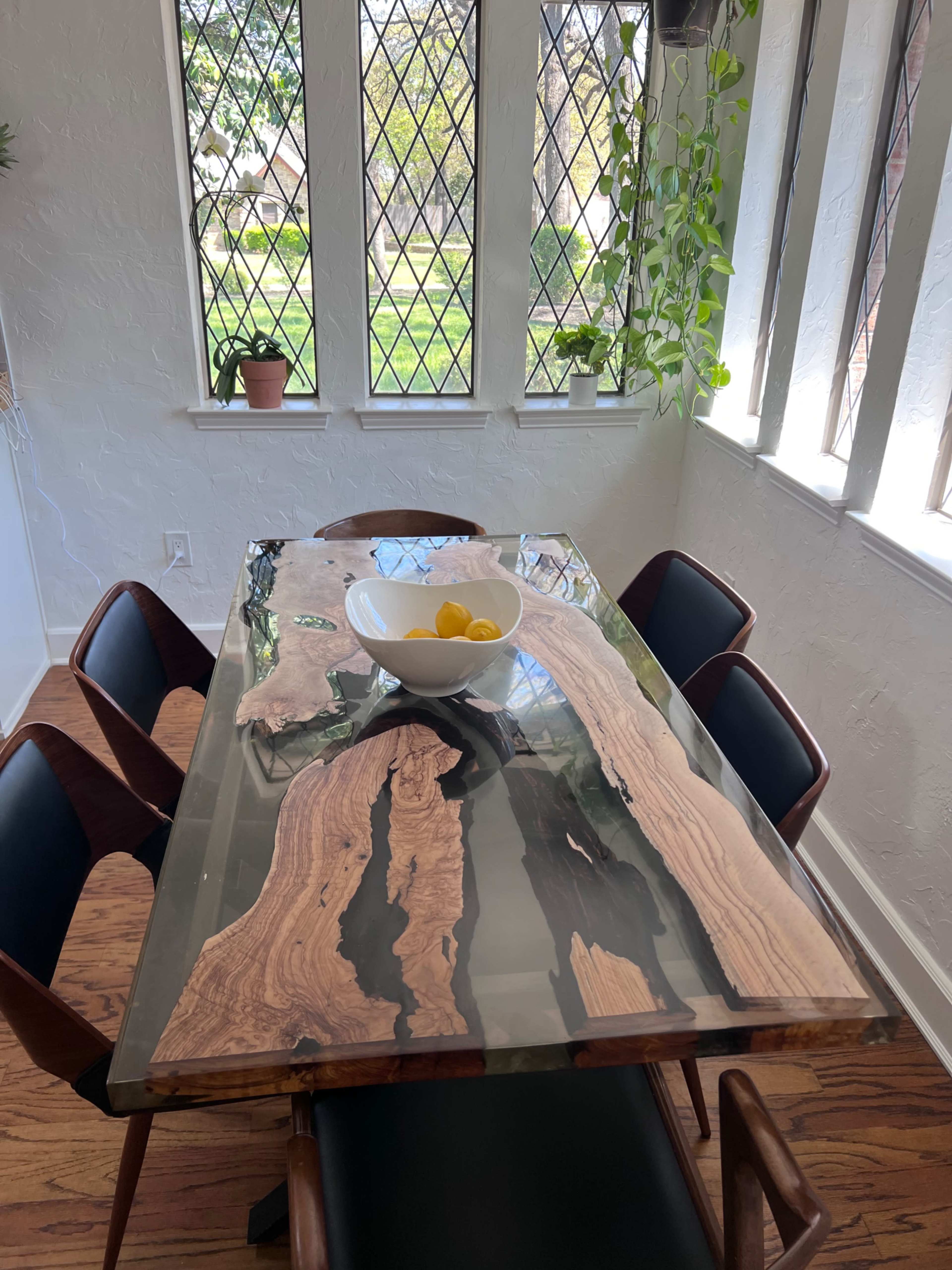 A wooden dining table with a glossy surface features a bowl of lemons and is surrounded by chairs in a sunlit room with large windows.