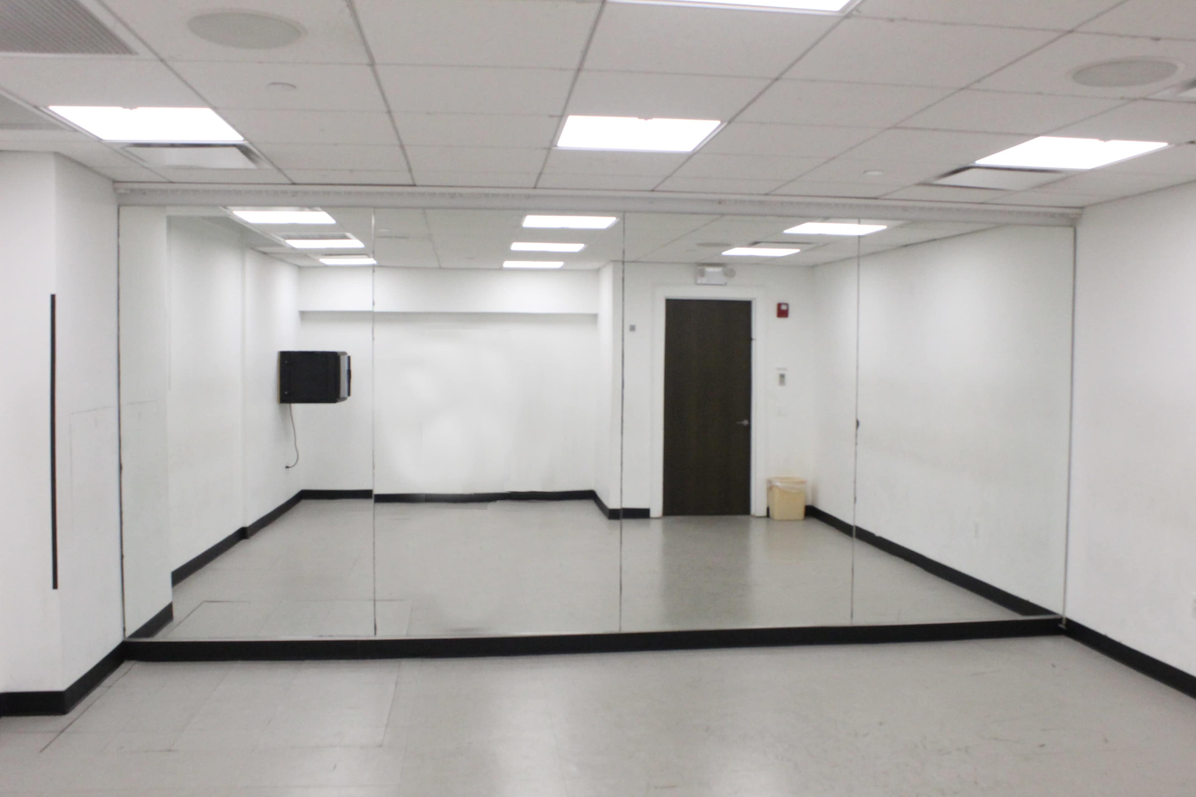 The image shows a simple, empty room with mirrors on one wall, fluorescent lights overhead, and a door on the right side.