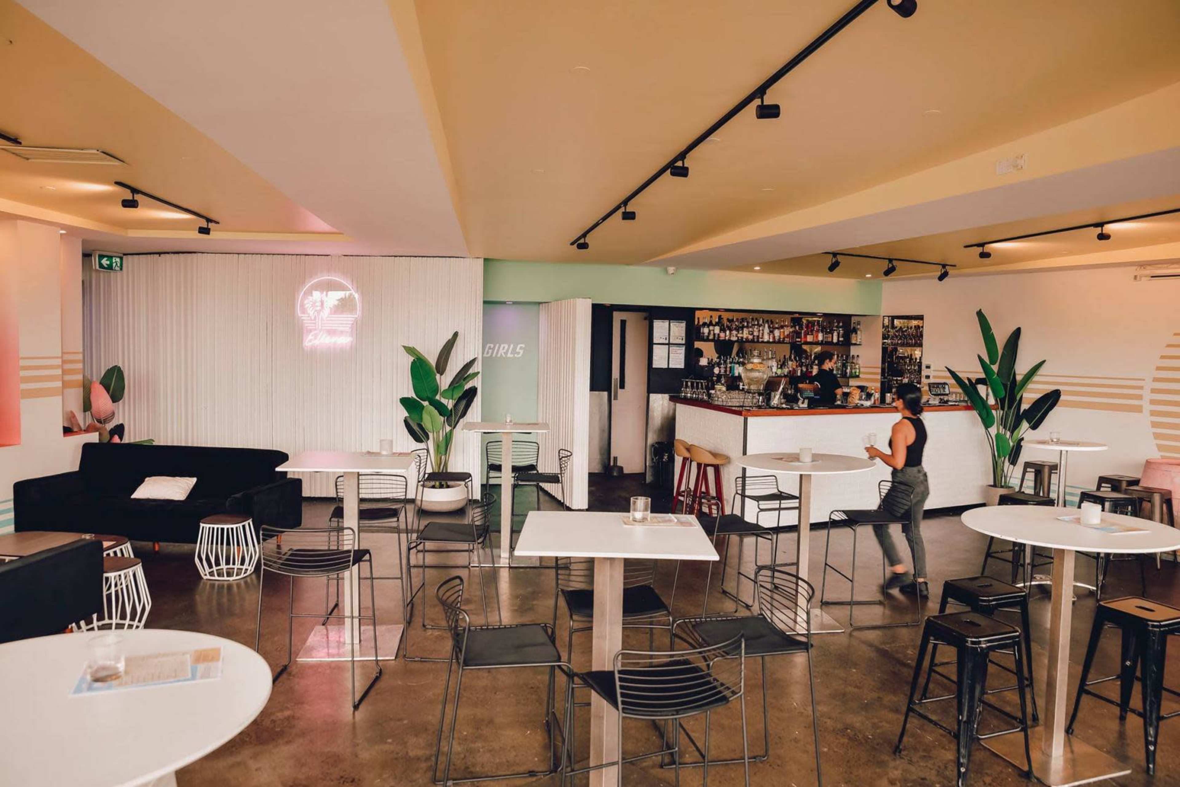 A spacious bar area features white tables and black seating, with a brightly lit counter and green plants set against a light-colored wall.