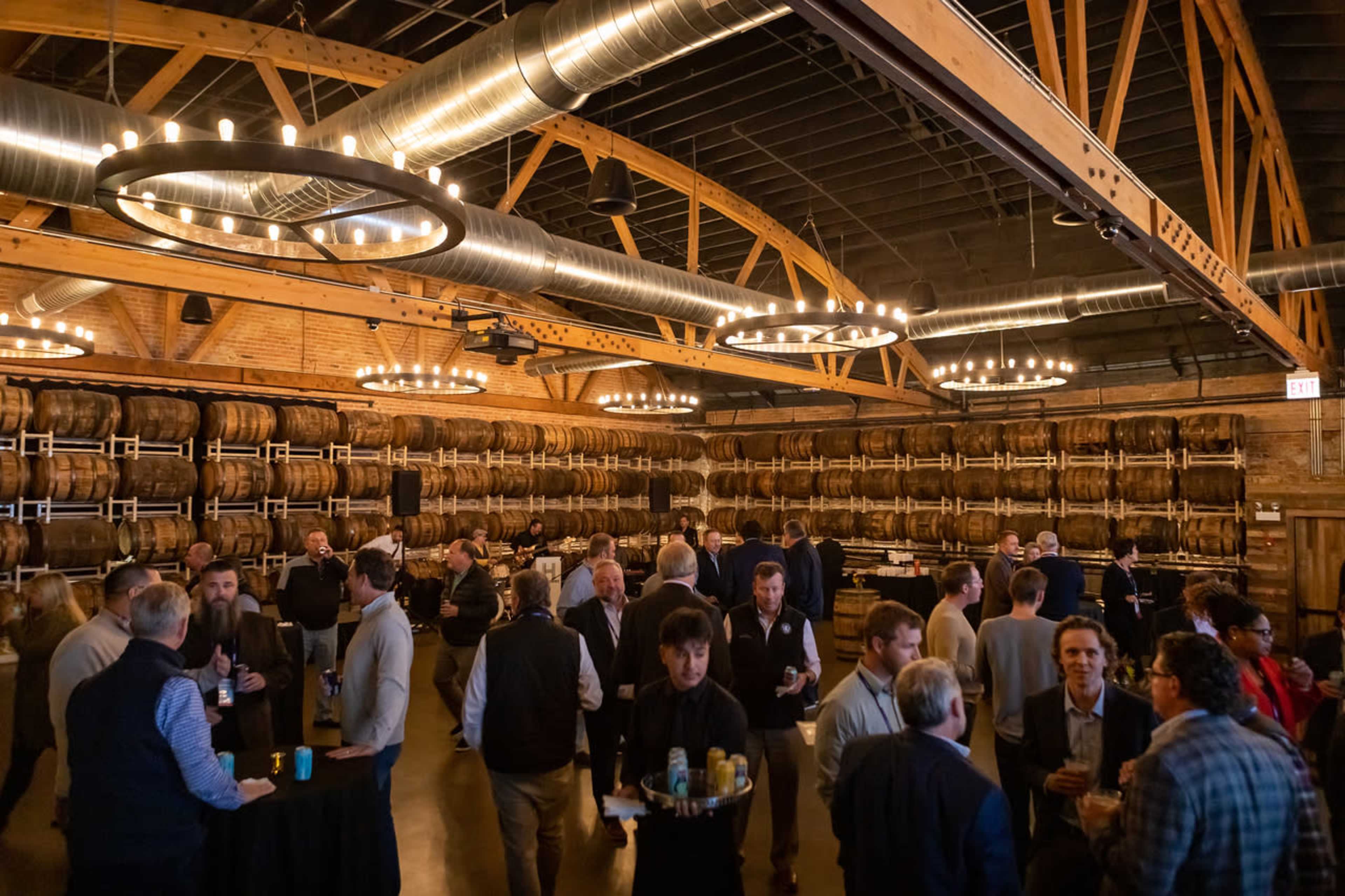 The image shows a spacious, rustic venue filled with people socializing among wooden barrels stacked against the walls.