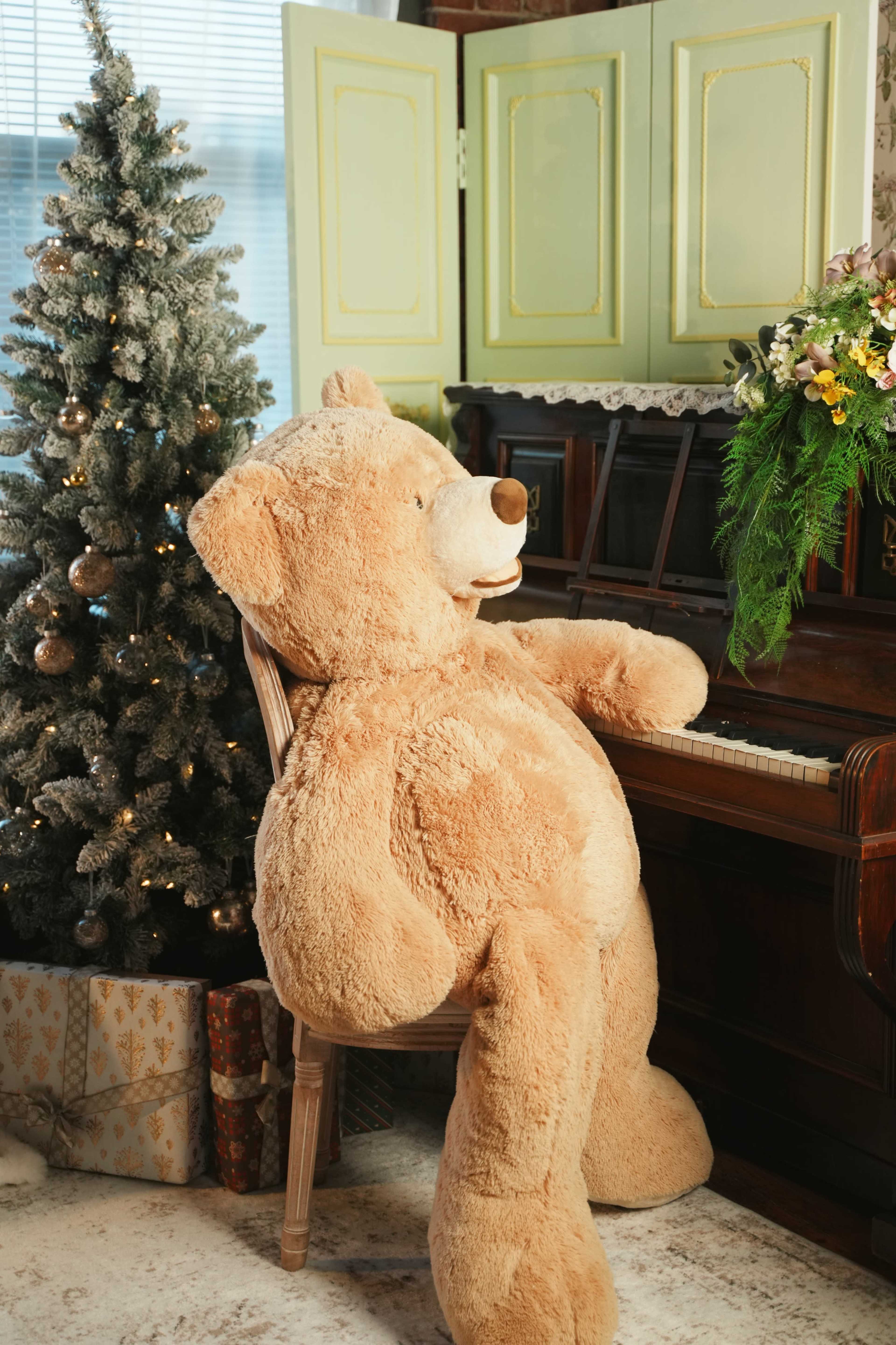 A large, plush teddy bear sits at a piano next to a decorated Christmas tree and festive gifts.