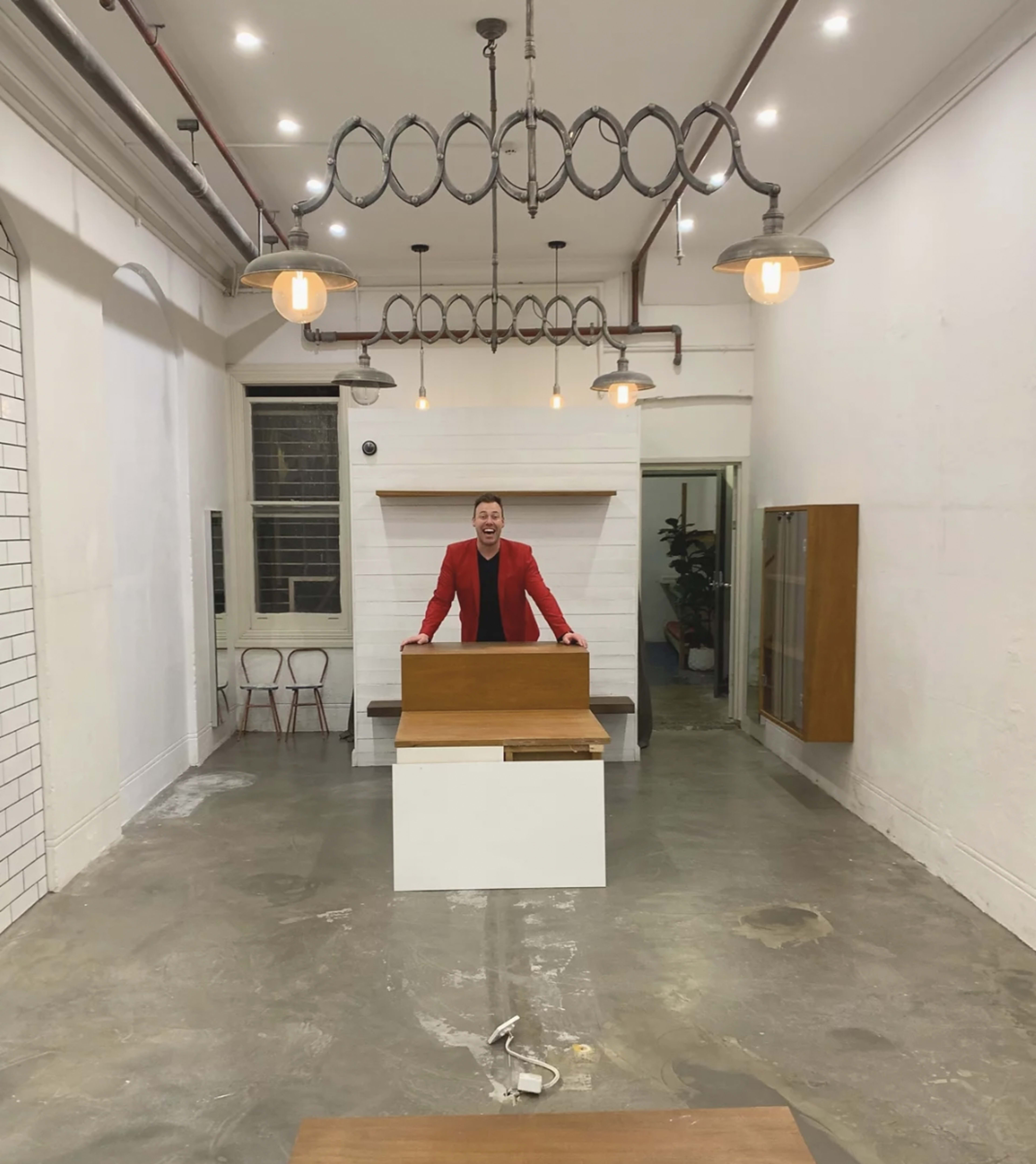 A man in a red blazer stands behind a table in a spacious, brightly lit room with industrial-style lighting and minimalistic decor.
