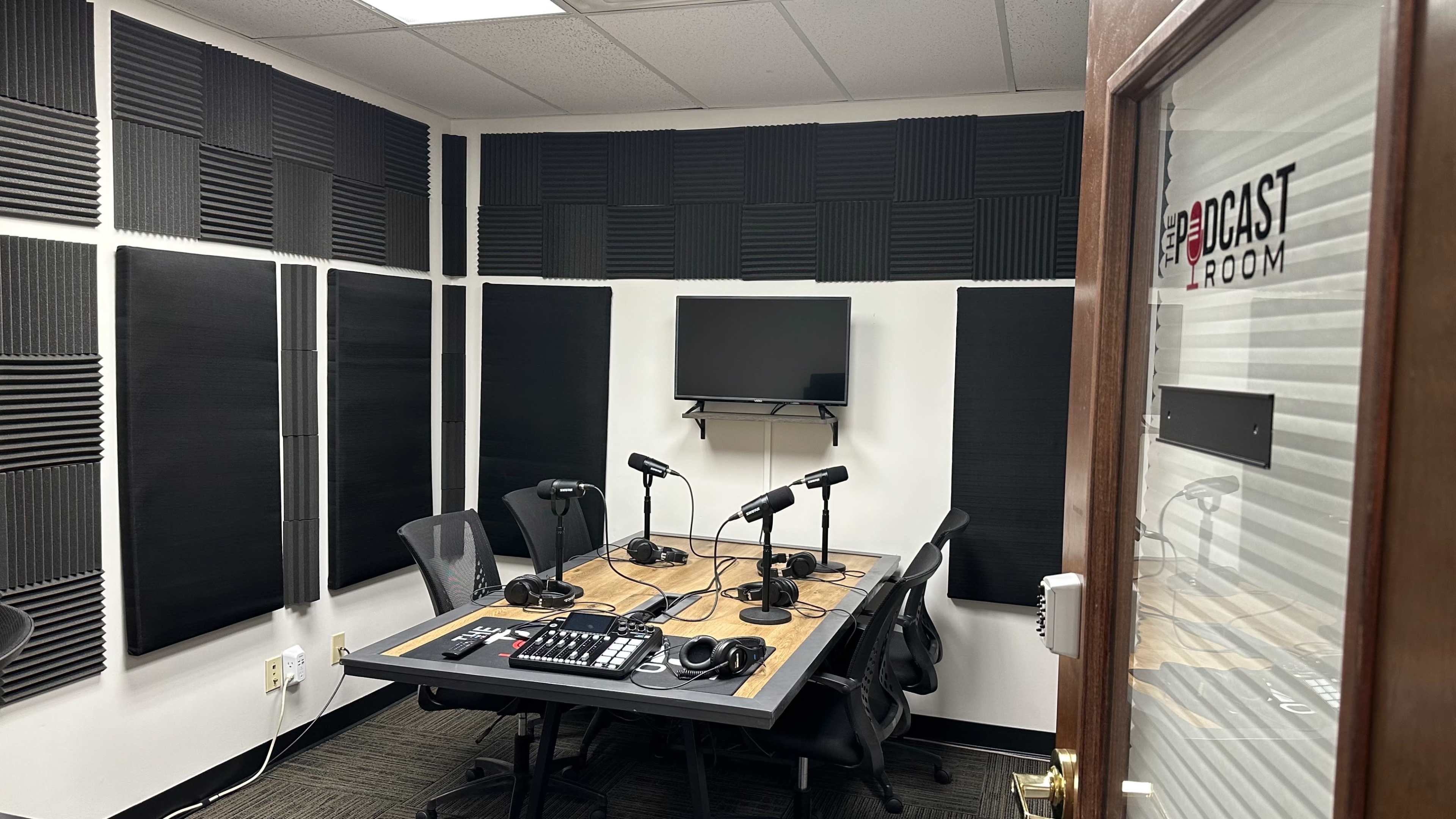 The image shows a podcast recording studio with soundproofing panels on the walls, a central table with microphones, and a television on the wall.