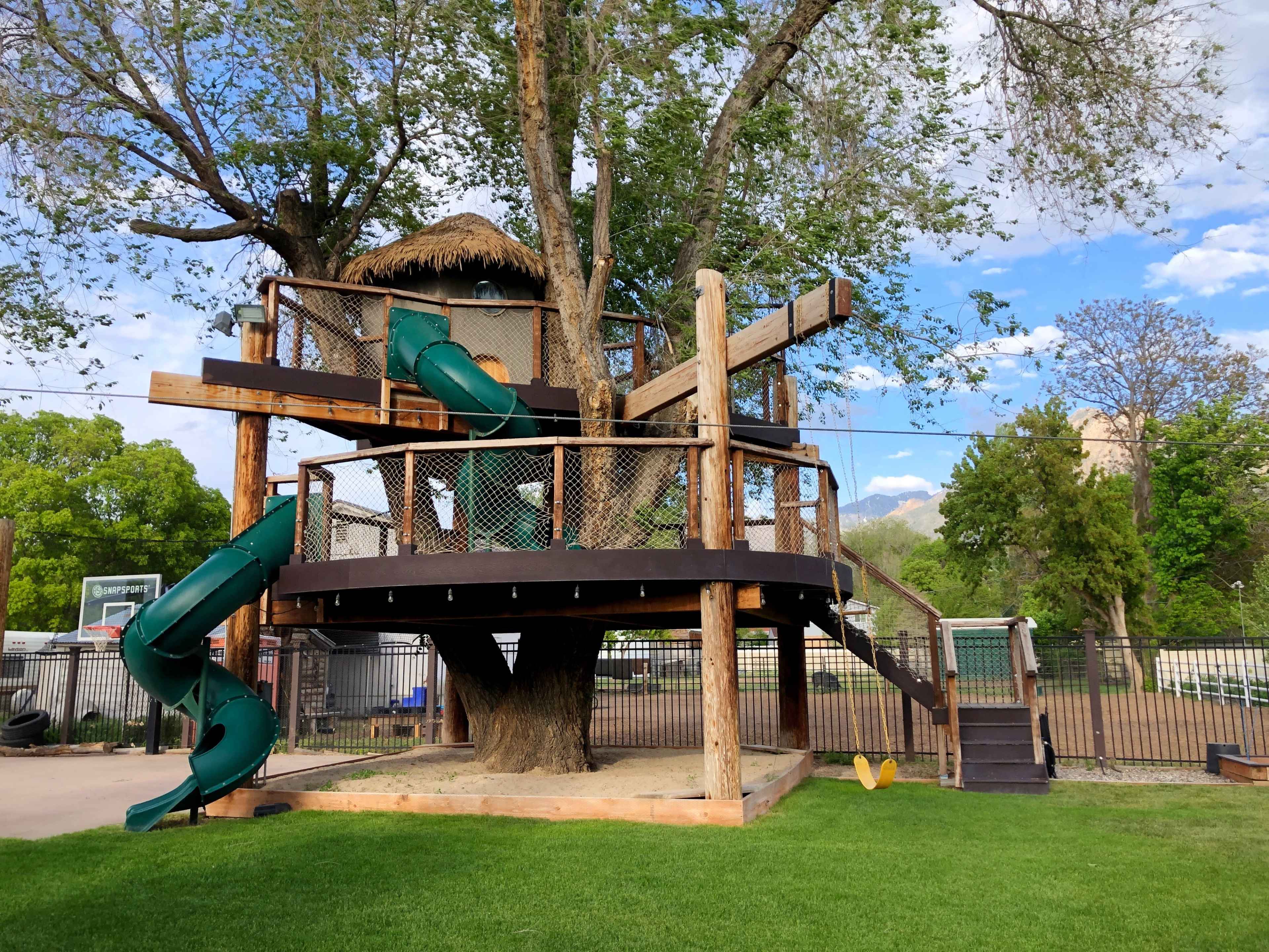 A large wooden treehouse with green slides, situated in a grassy area surrounded by trees and a fence.
