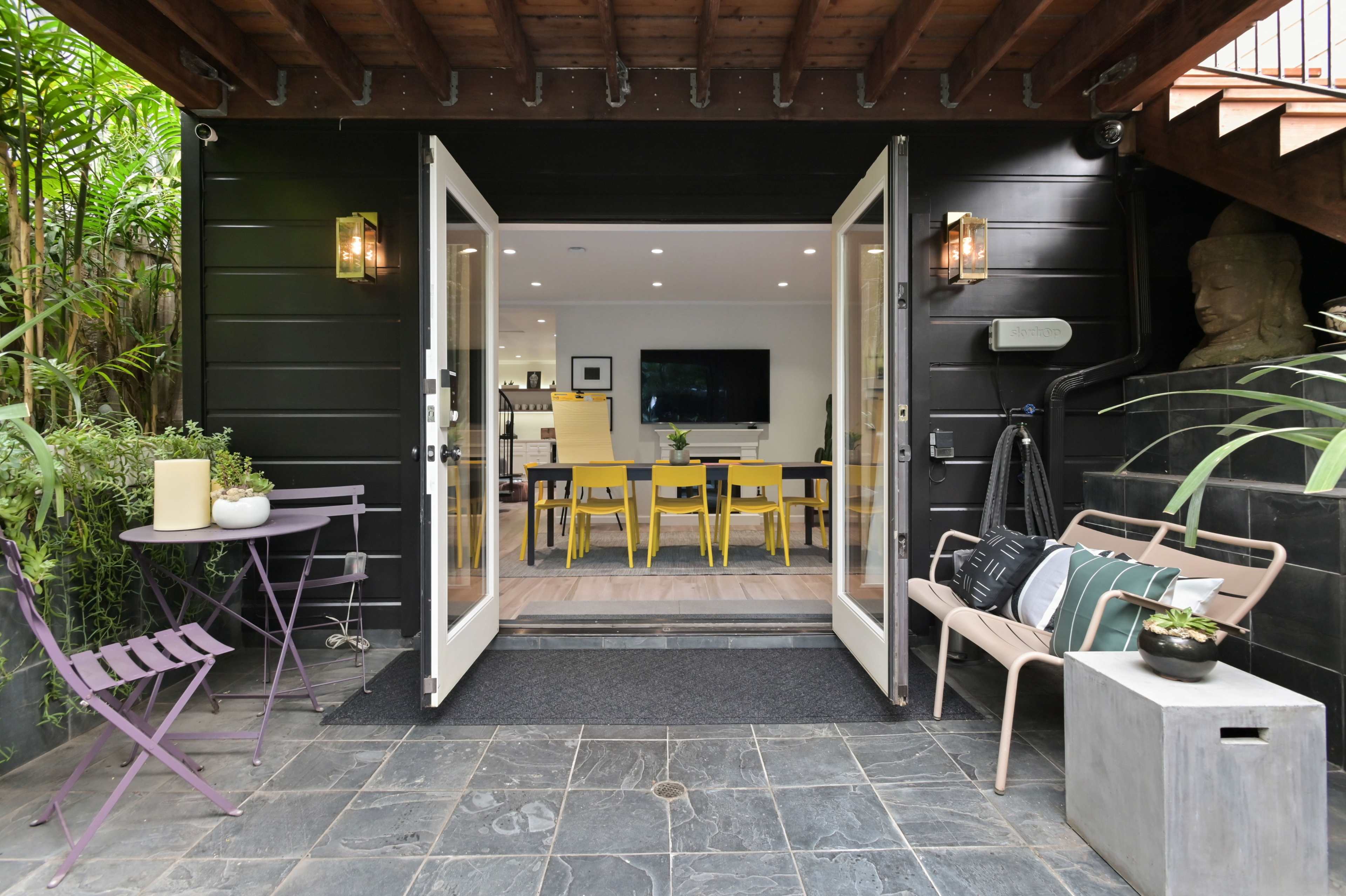 A set of double doors open into a modern dining area with yellow chairs, surrounded by greenery and natural stone features.