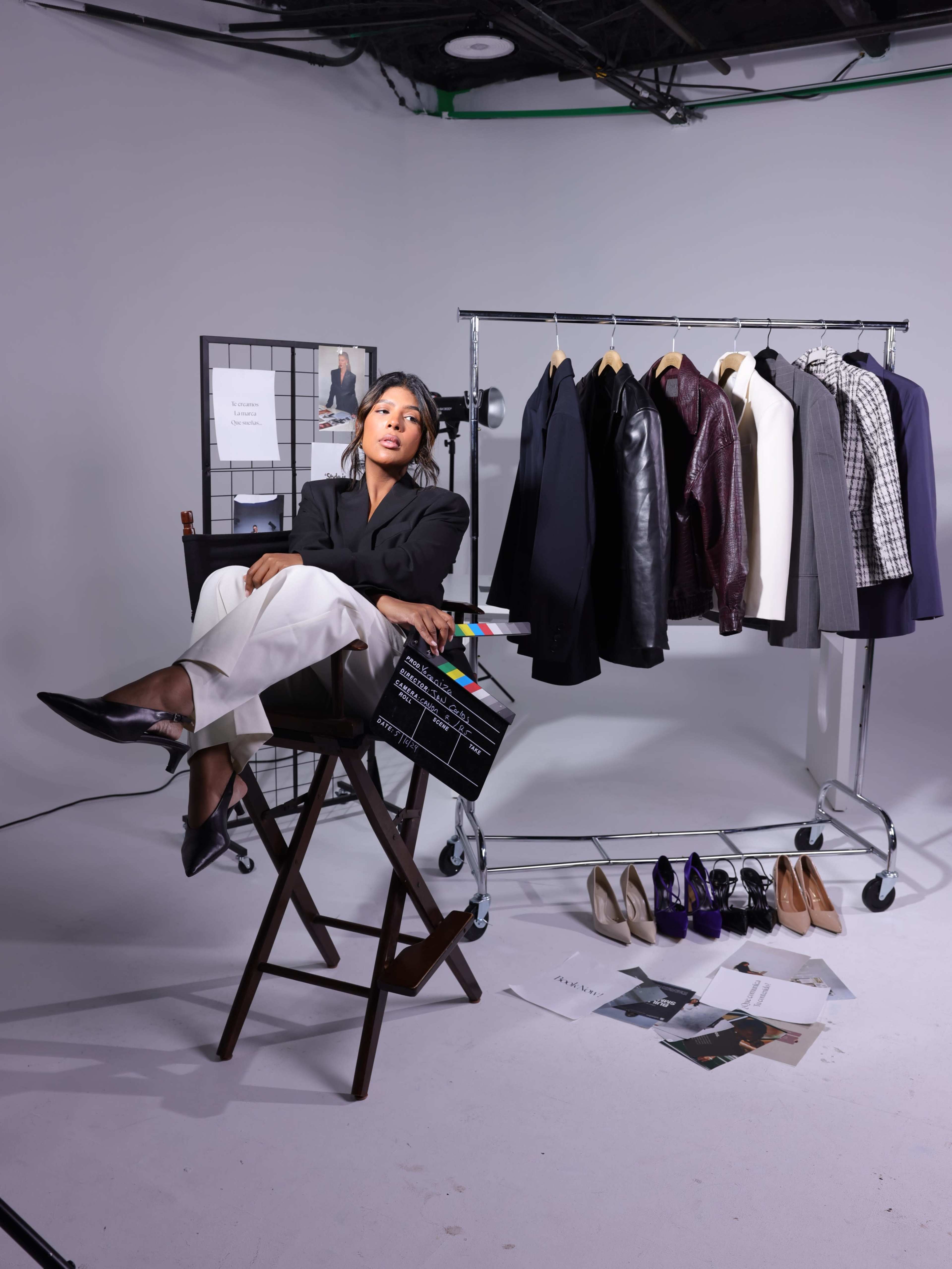 A woman sits on a director's chair holding a clapboard in front of a clothing rack displaying various outfits and a selection of shoes on the floor.