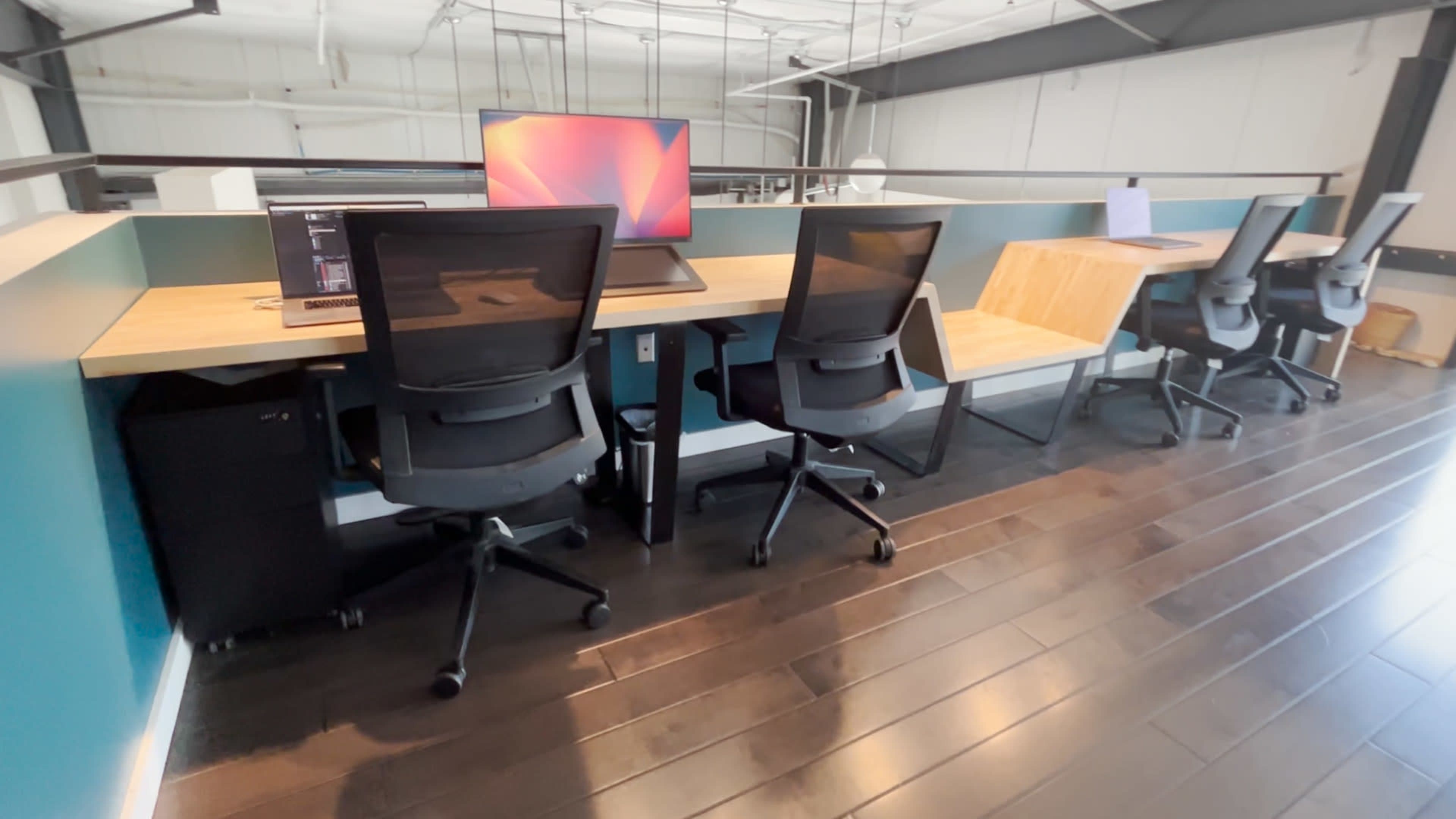 A row of modern office desks equipped with computer monitors and ergonomic chairs is set against a teal wall in a spacious workspace.