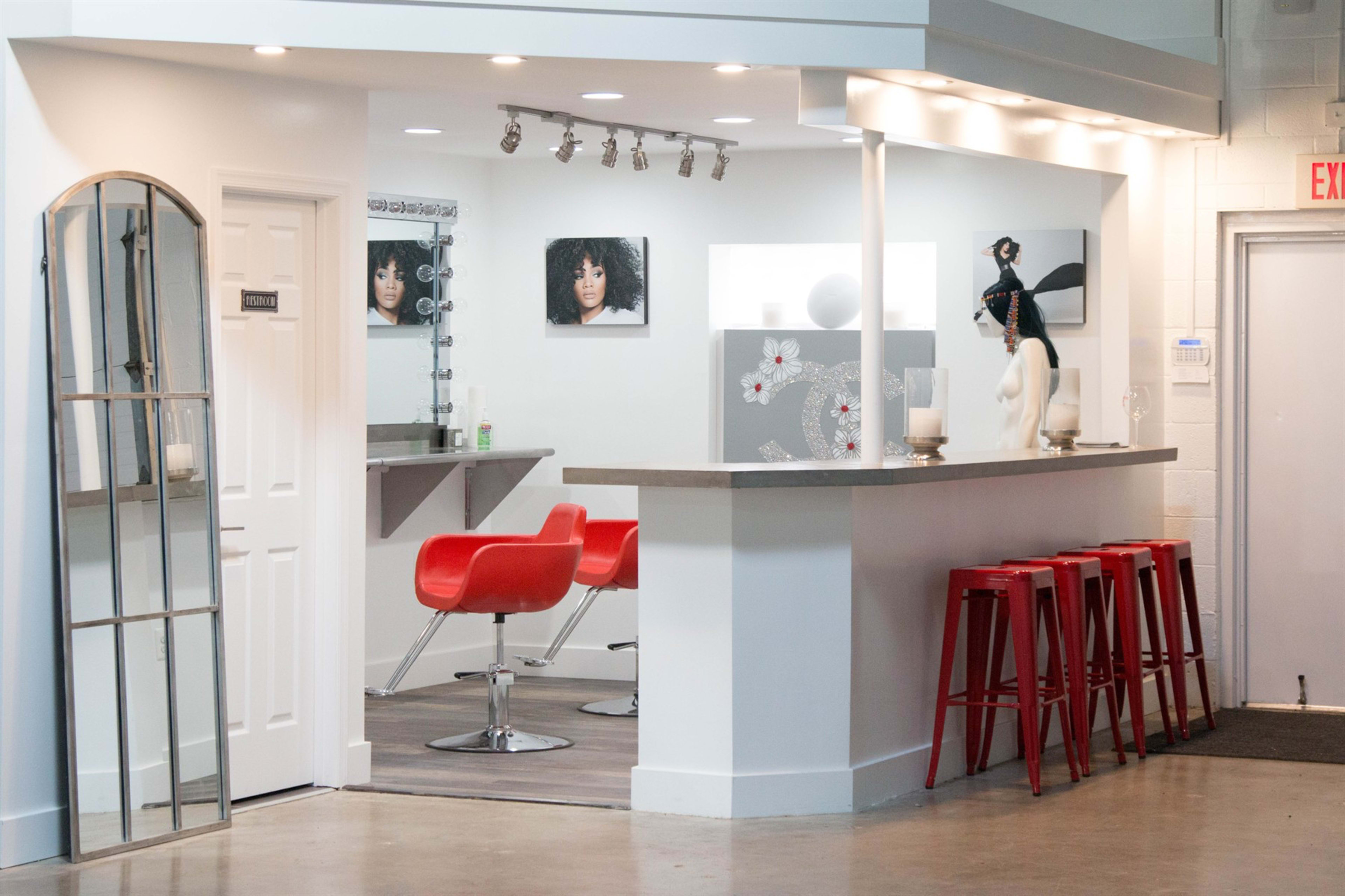 A modern salon interior features a reception area with a sleek counter, red bar stools, and mirrors alongside stylish wall art.