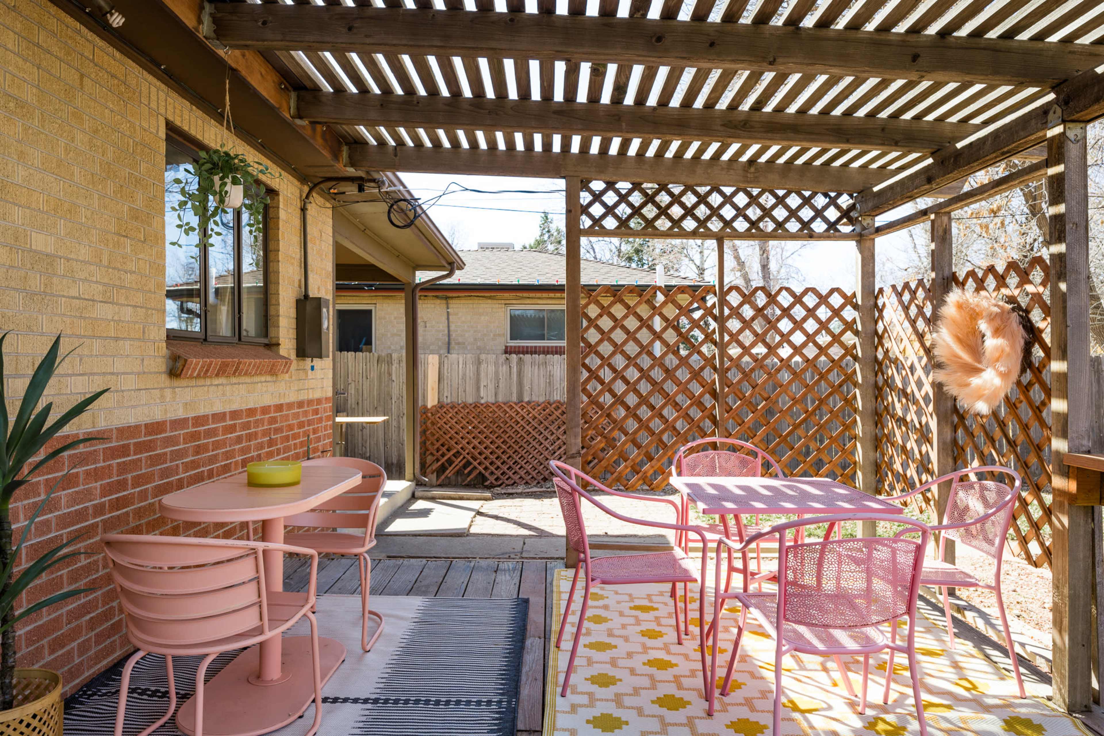 The image shows a patio area with a wooden pergola, featuring a pink table and chairs, and a patterned yellow and white rug.