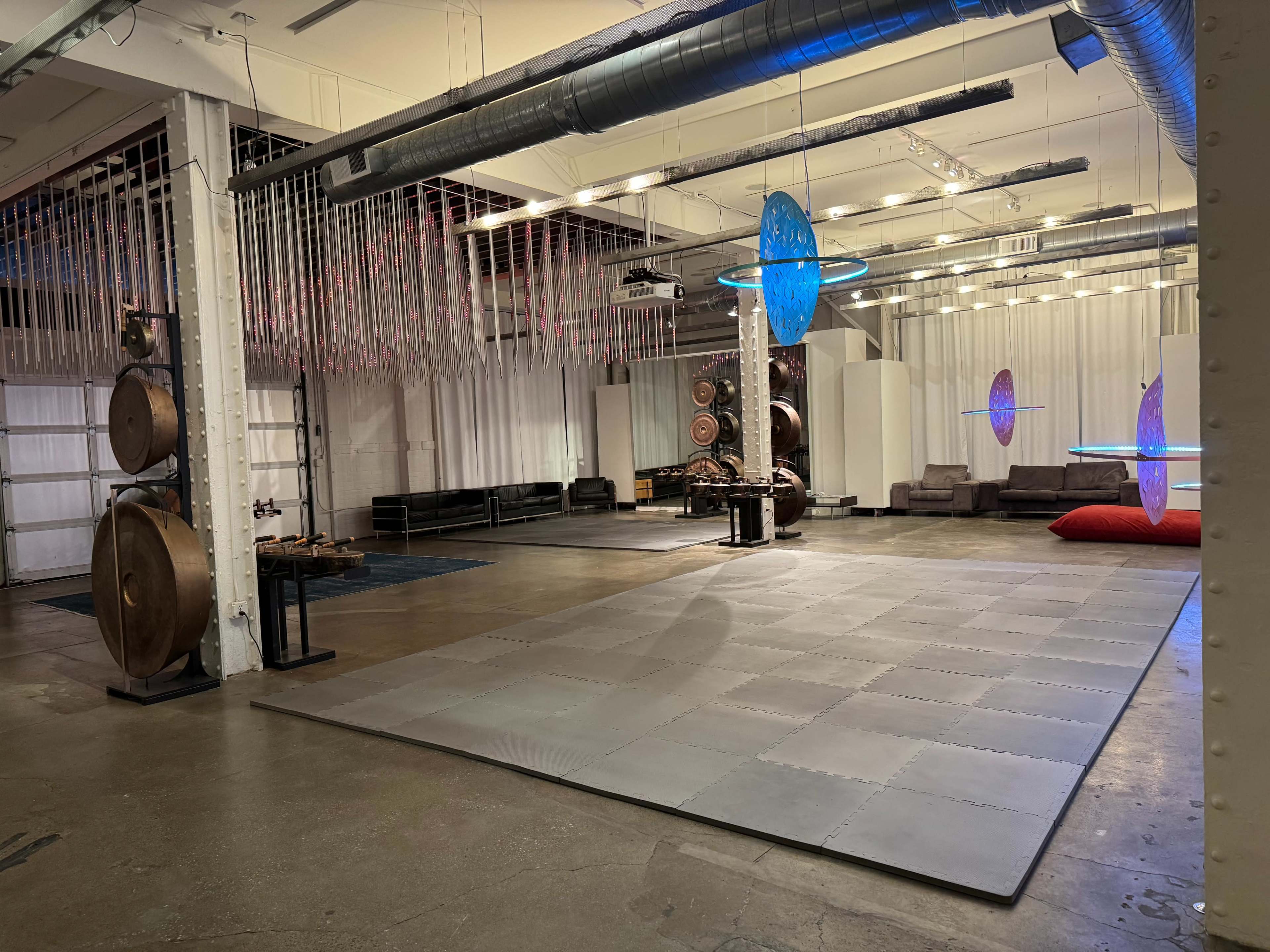 The image shows an empty industrial space with gray flooring, circular art pieces hanging from the ceiling, and various seating arrangements along the walls.