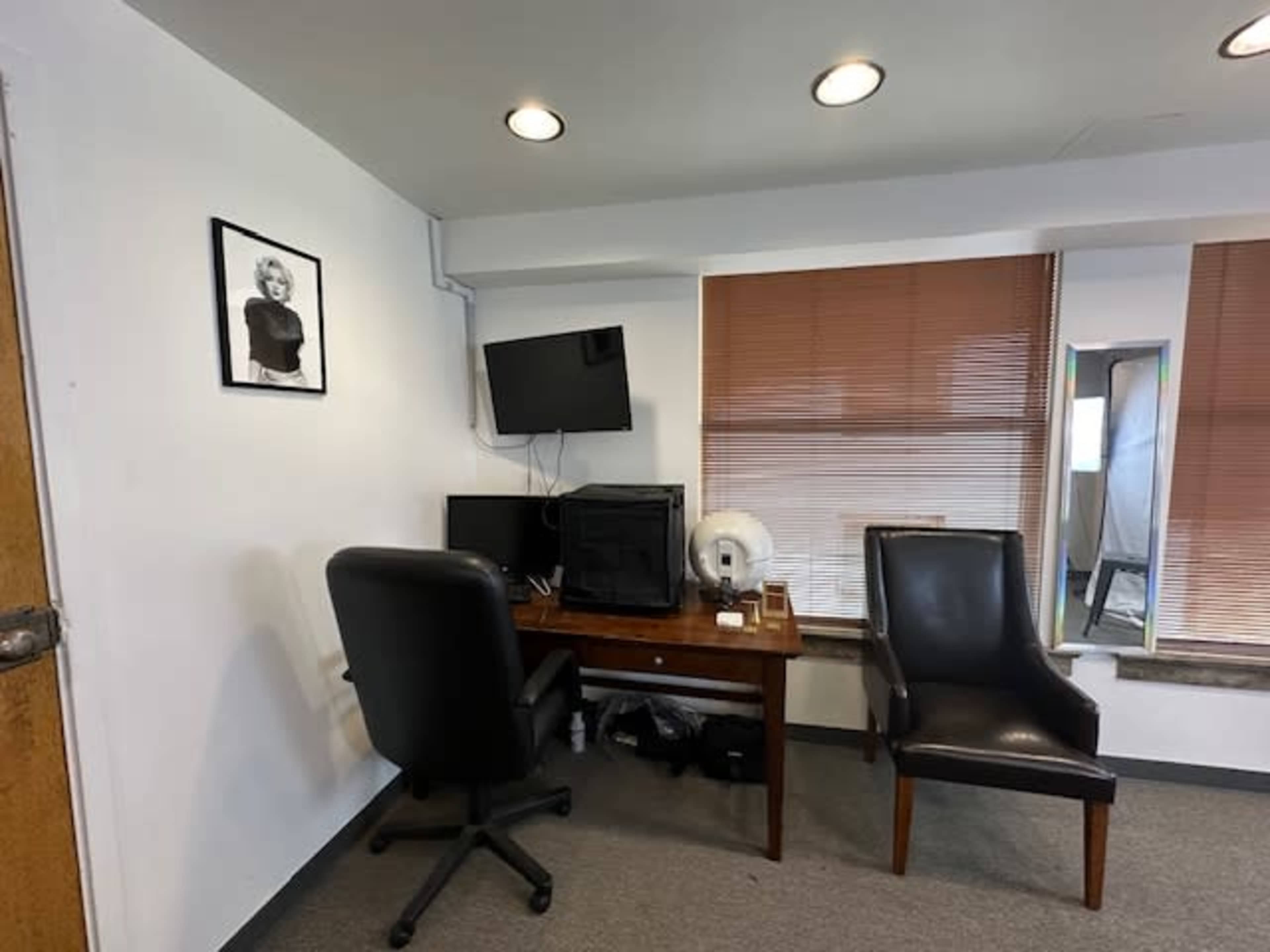 The image shows a small office space with a desk, two chairs, a television mounted on the wall, and window blinds partially raised.