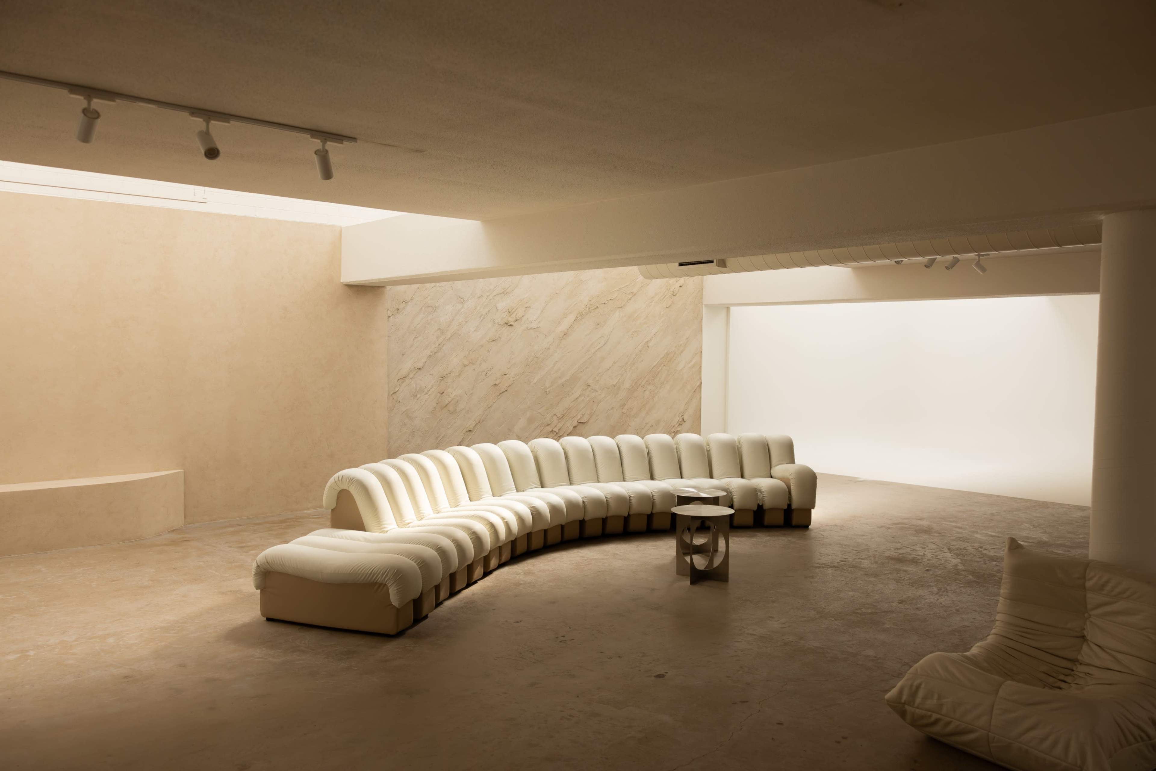 The image shows a modern, curved white sectional sofa placed in a minimalist interior space with neutral-toned walls and a plain backdrop.