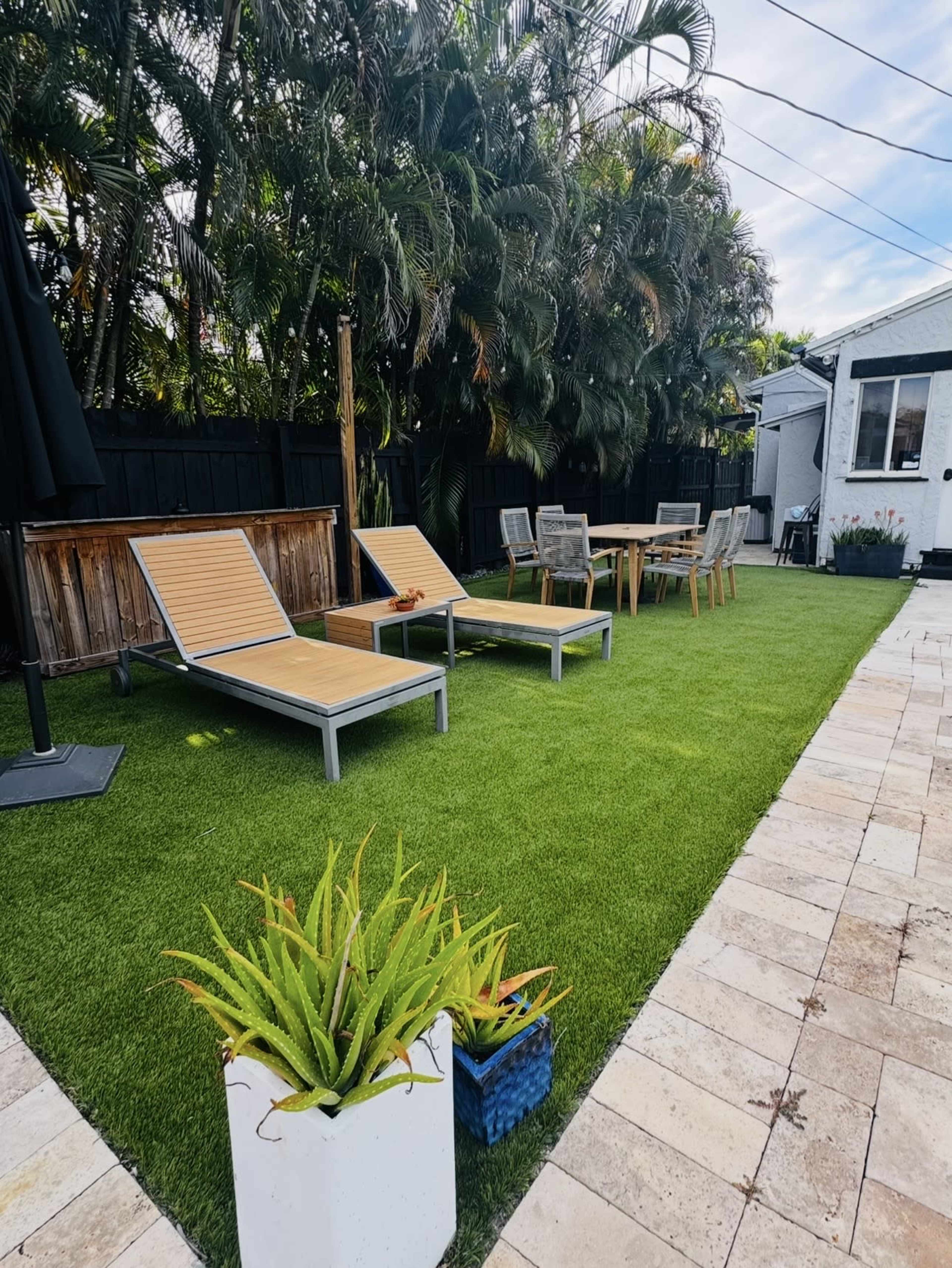 The image shows a well-maintained backyard featuring two lounge chairs, a dining table with chairs, and a planter, all surrounded by artificial grass and tropical plants.