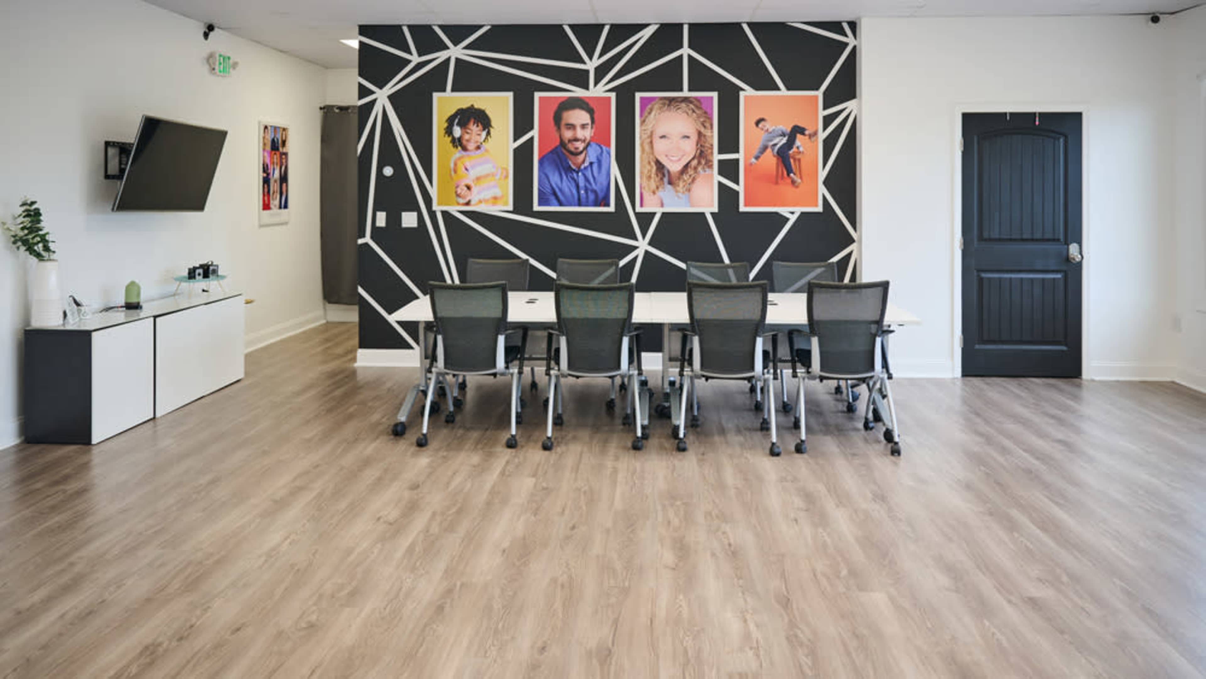 The image shows a modern conference room featuring a long table with chairs arranged around it and a wall decorated with framed portraits.