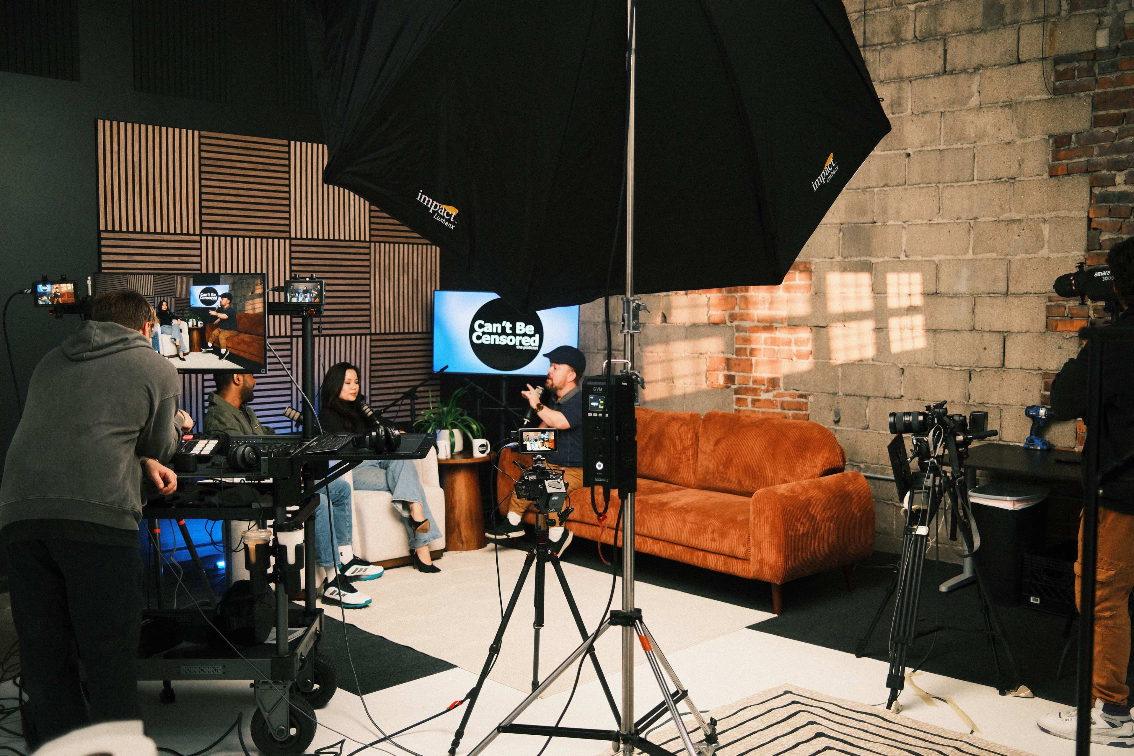 A studio setup features a discussion between two people seated in front of a camera, with multiple filming equipment and a large umbrella light.