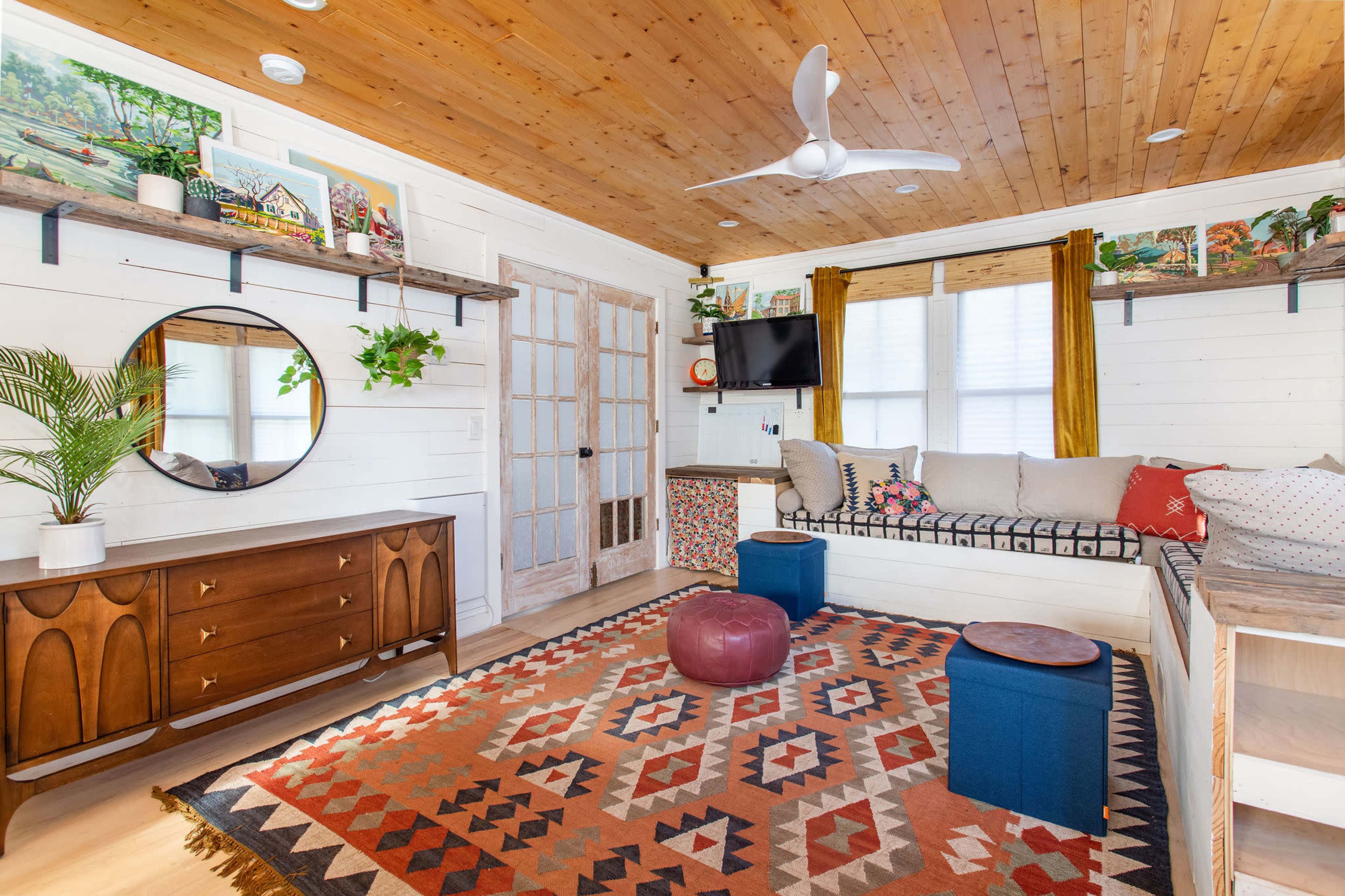 A bright living room with wooden paneling, a ceiling fan, and a mix of modern and traditional furnishings, including a large area rug, a wall-mounted TV, and a cozy seating area.
