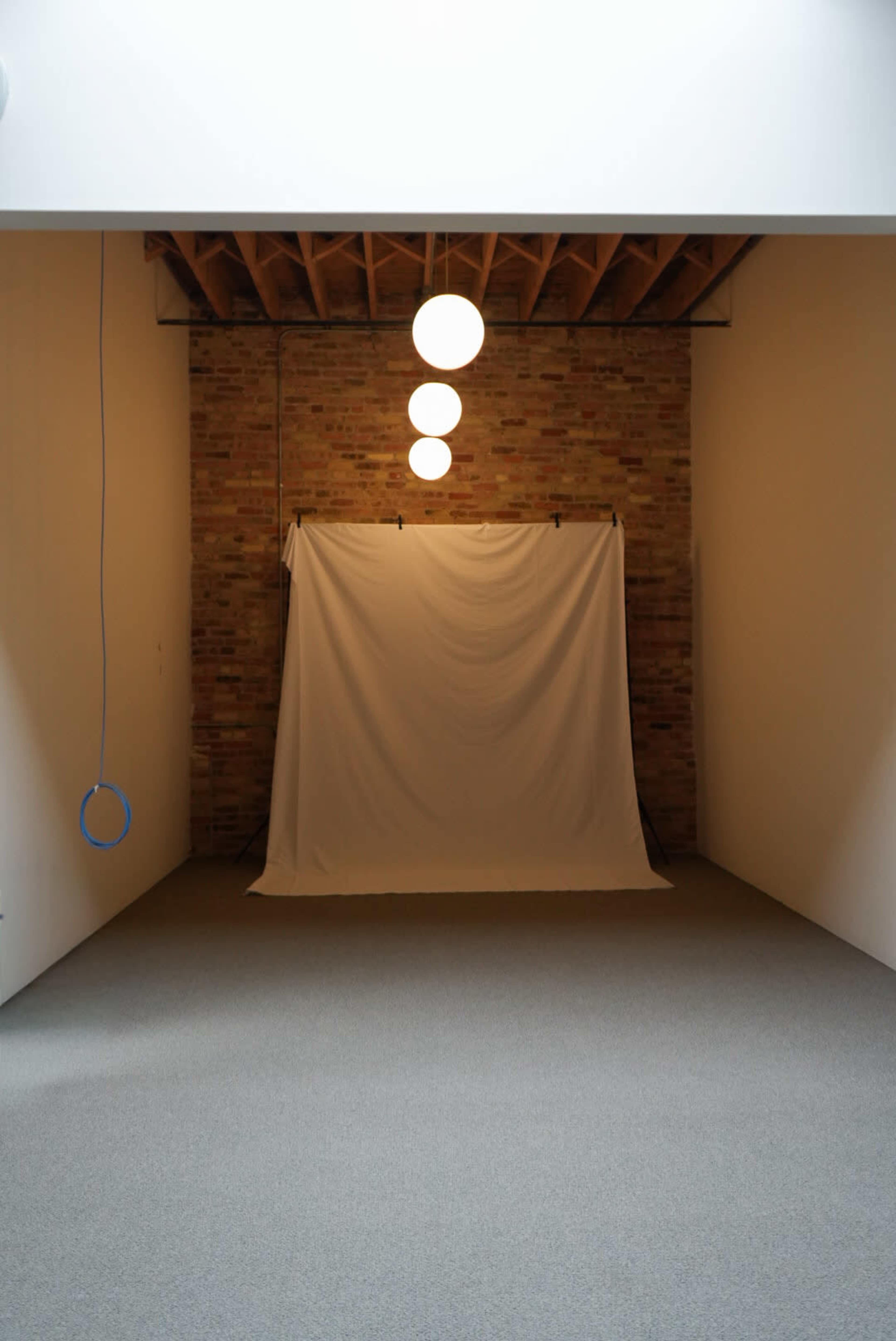 The image shows an empty indoor studio space with a white backdrop, exposed brick walls, and four round pendant lights hanging from the ceiling.