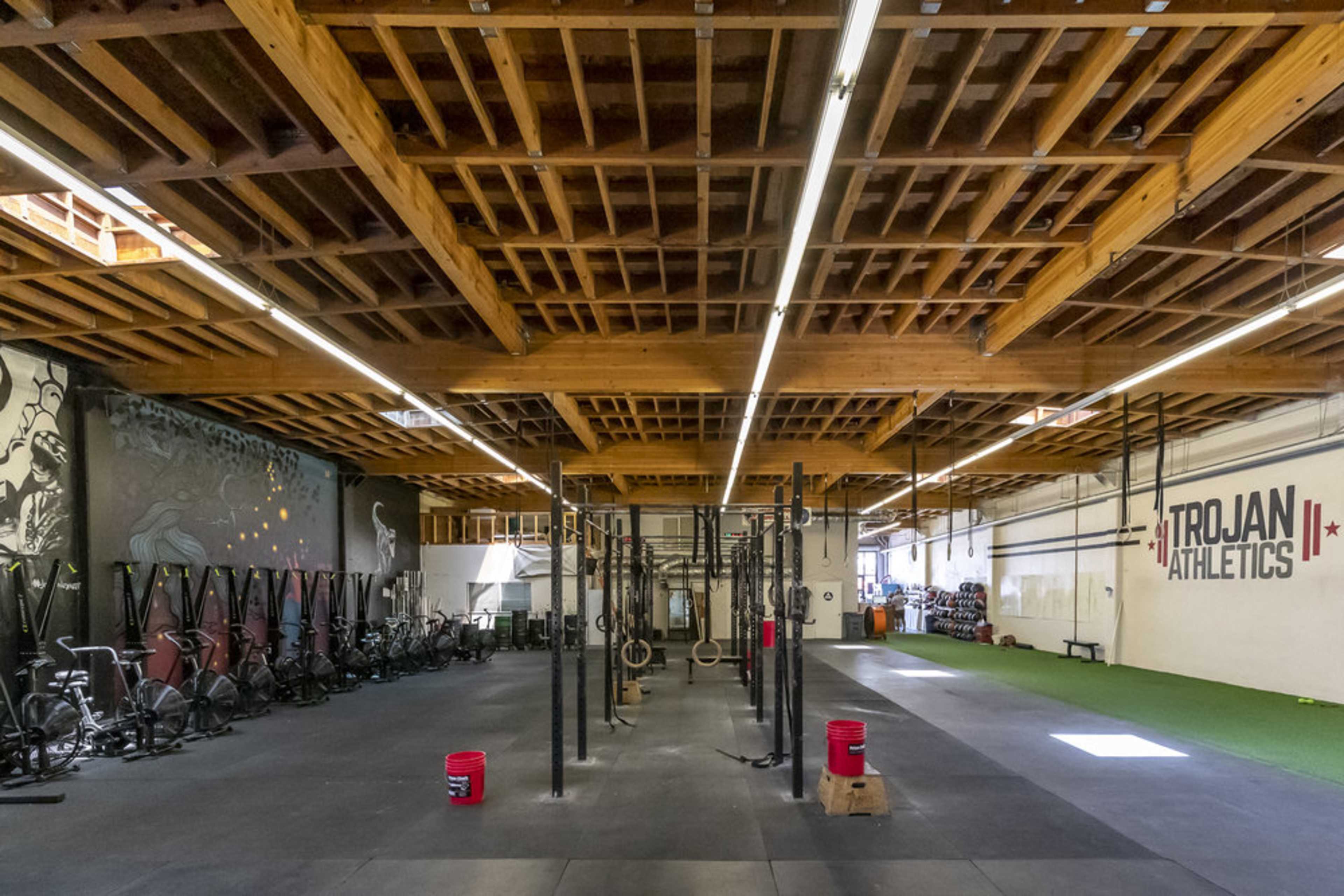 The image shows a spacious gym interior with wooden beams, workout equipment arranged along the walls, and designated exercise areas with pull-up bars and exercise mats.