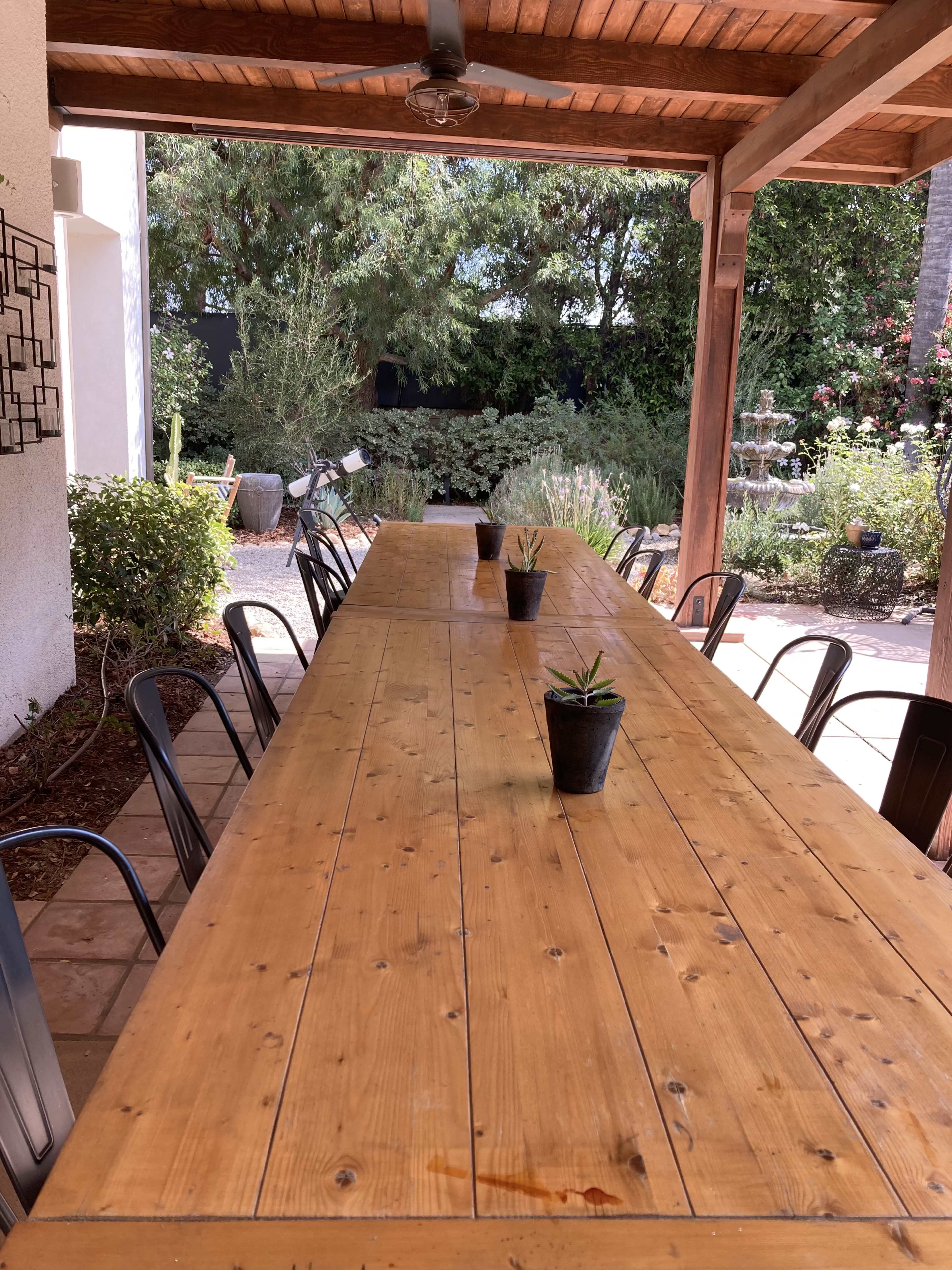 A long wooden dining table with potted plants is set under a covered patio surrounded by greenery.