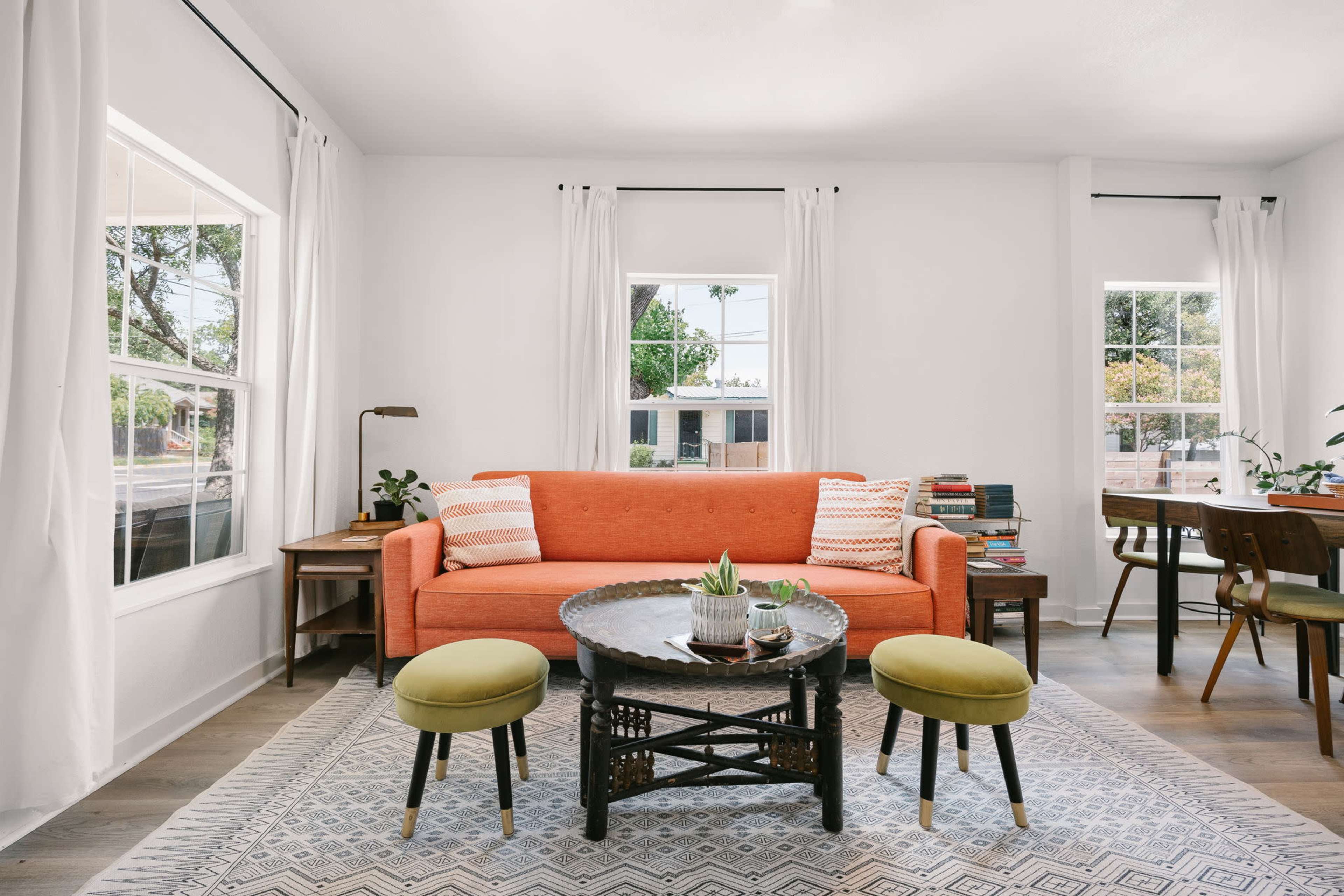 The image depicts a bright living room featuring an orange couch, a round coffee table, and two green ottomans, with large windows allowing natural light to enter.