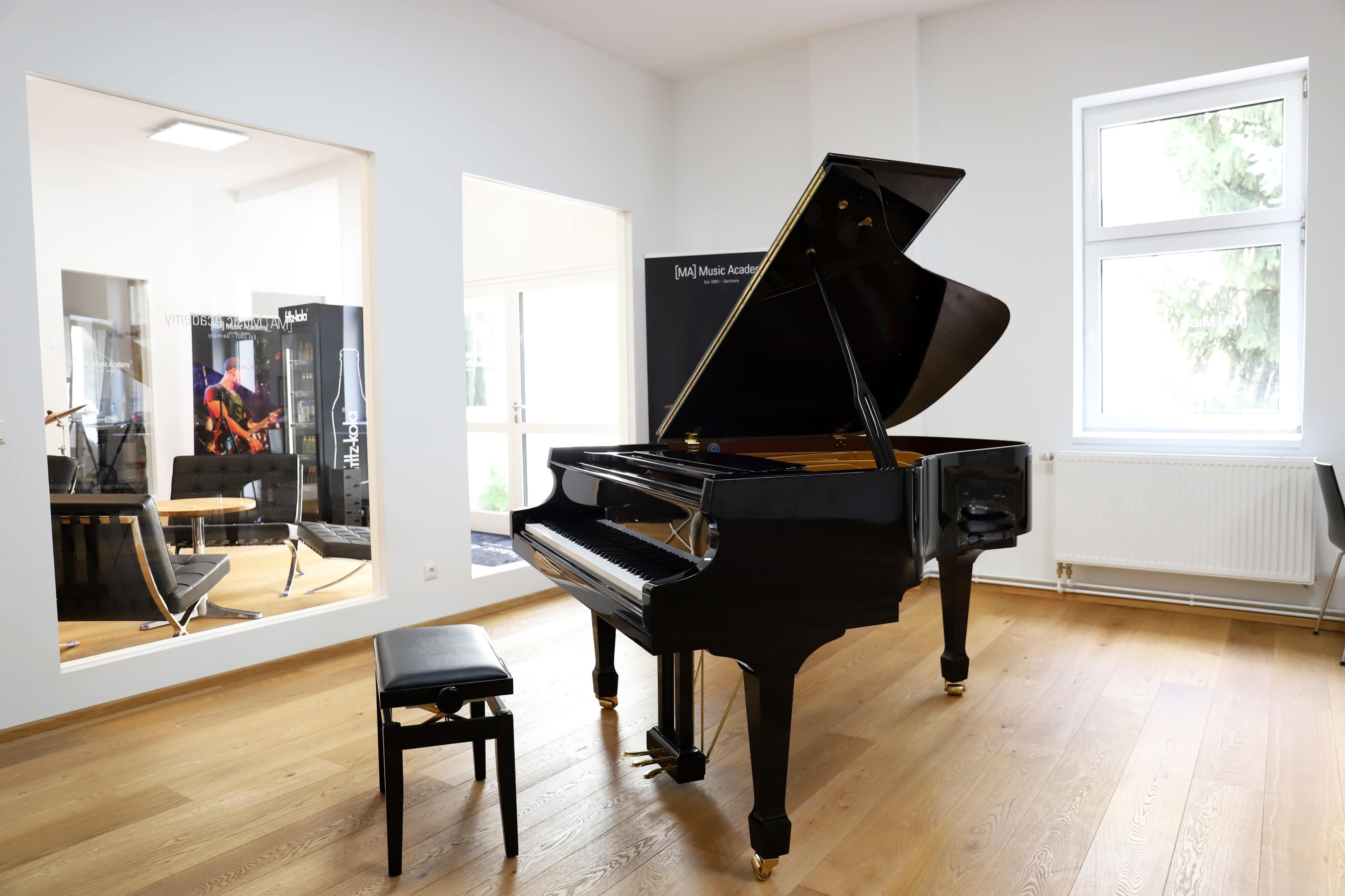 A black grand piano with a matching bench is situated in a bright room featuring wooden floors and large windows.