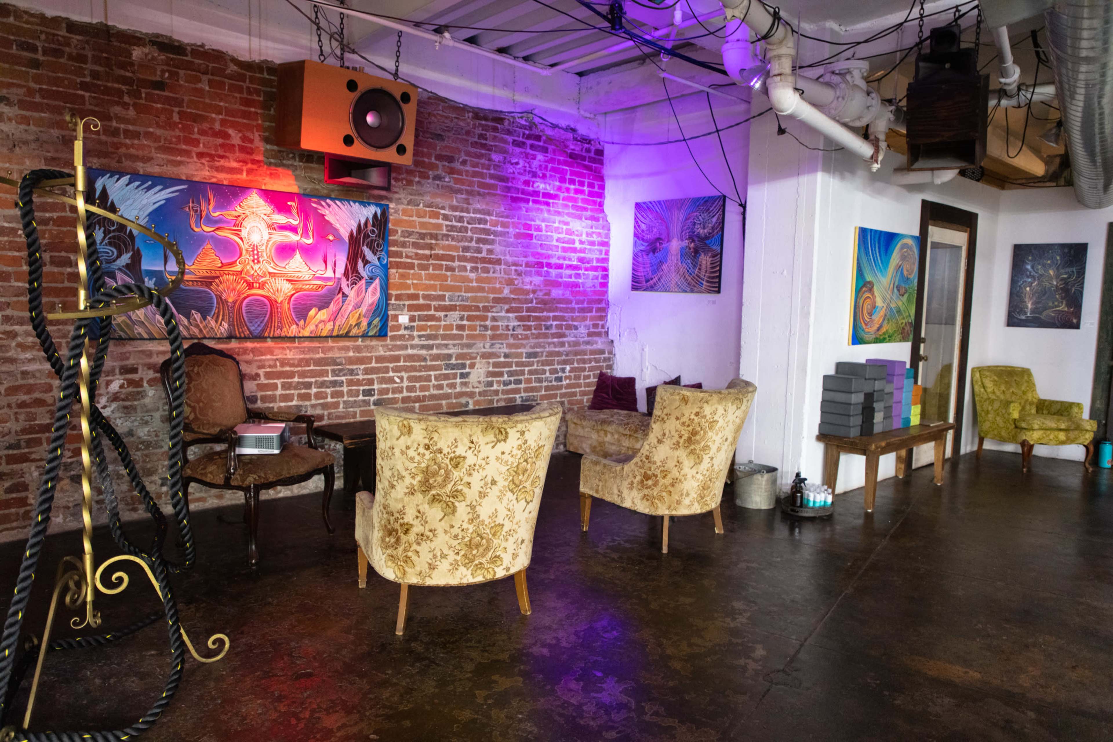 A spacious room with exposed brick walls, featuring two patterned chairs, colorful artworks, and a few stacks of boxes in the corner.
