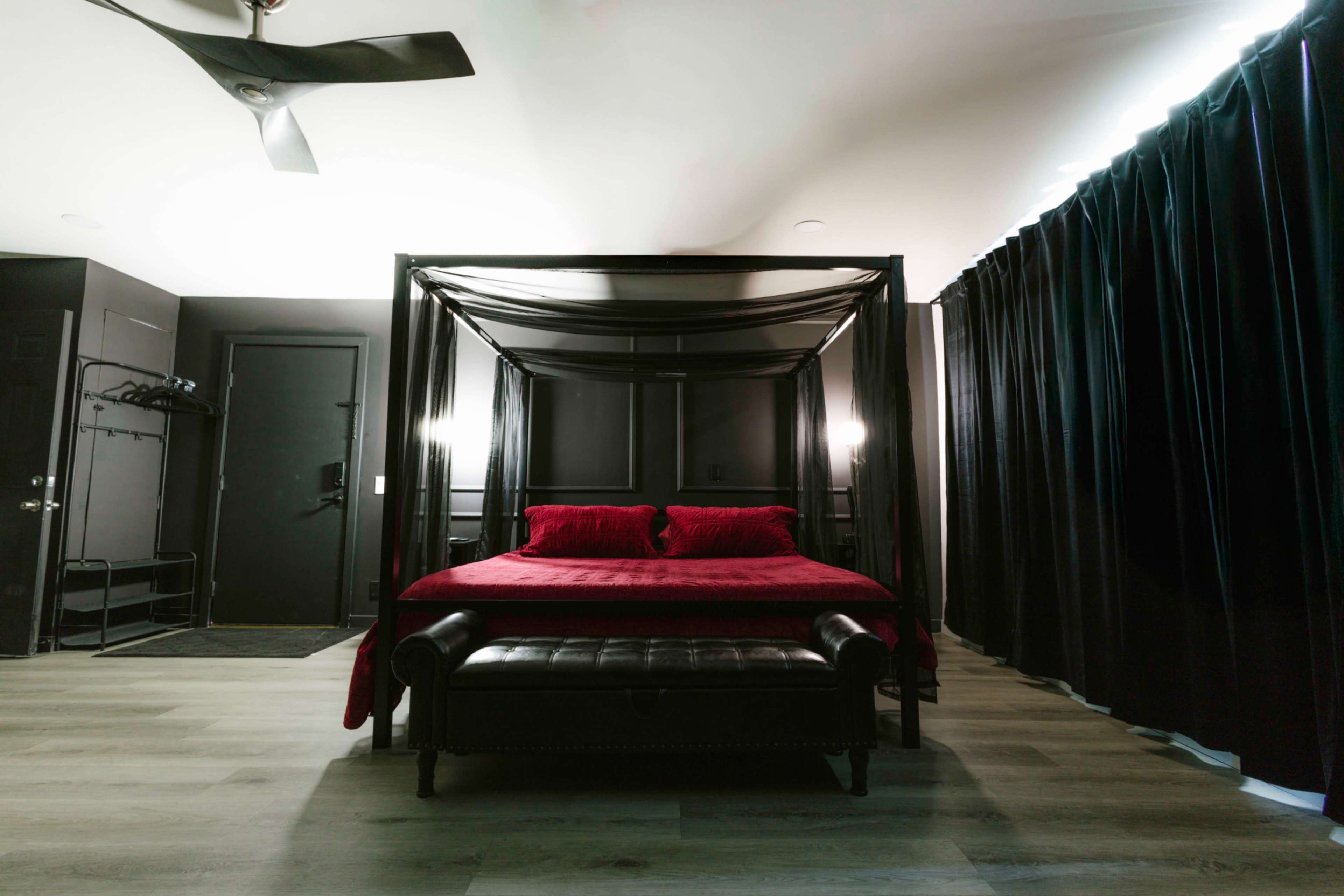 A black four-poster bed with red bedding is positioned in a dimly lit room featuring dark walls and a curtain-covered window.
