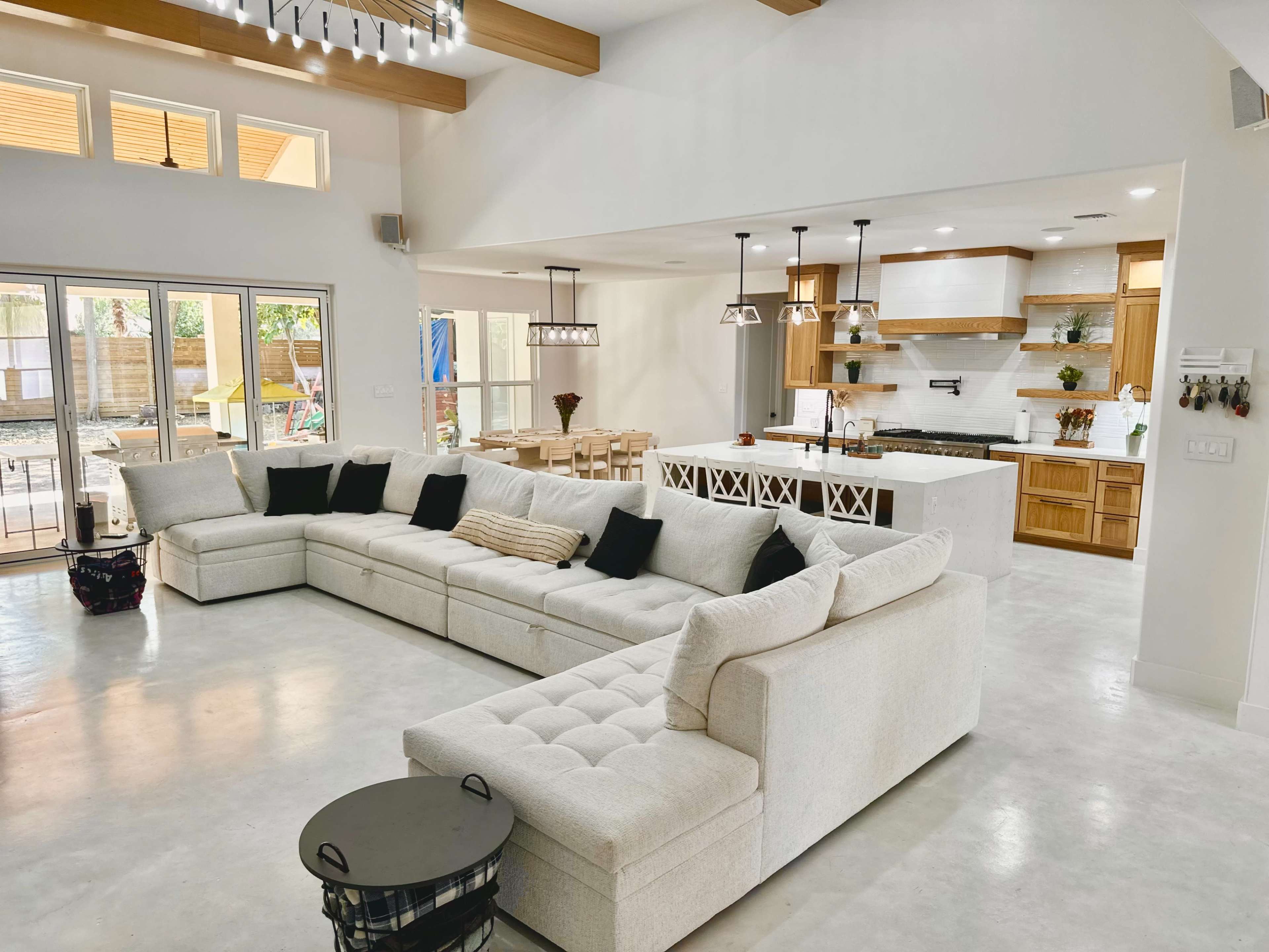 The image shows a spacious, modern living area featuring a large sectional sofa, a dining area with wooden chairs, and an open kitchen with wooden cabinets and a central island.