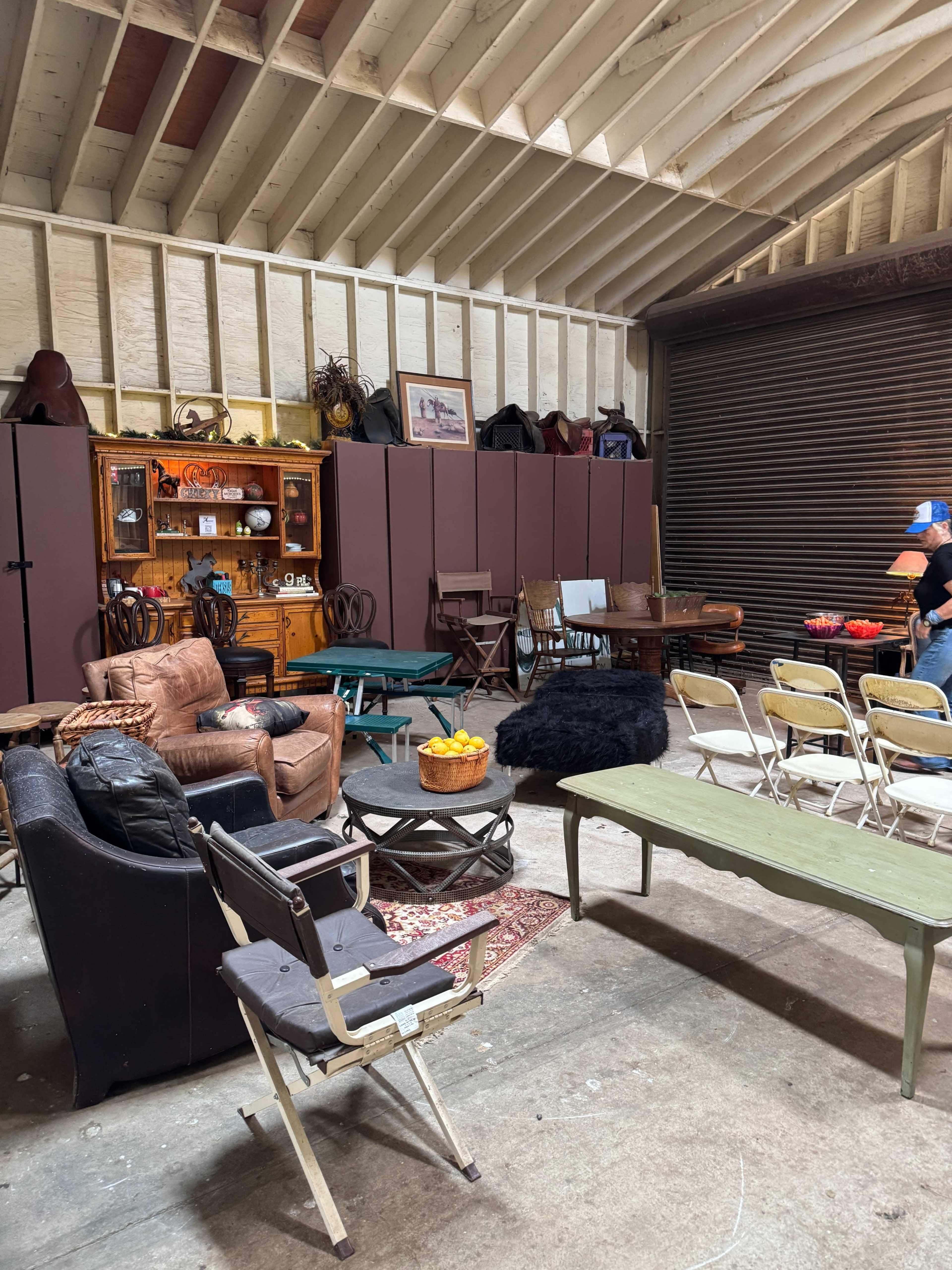 The image shows a room filled with various pieces of furniture, including chairs, tables, and a cabinet, arranged in a storage space with wooden beams overhead.