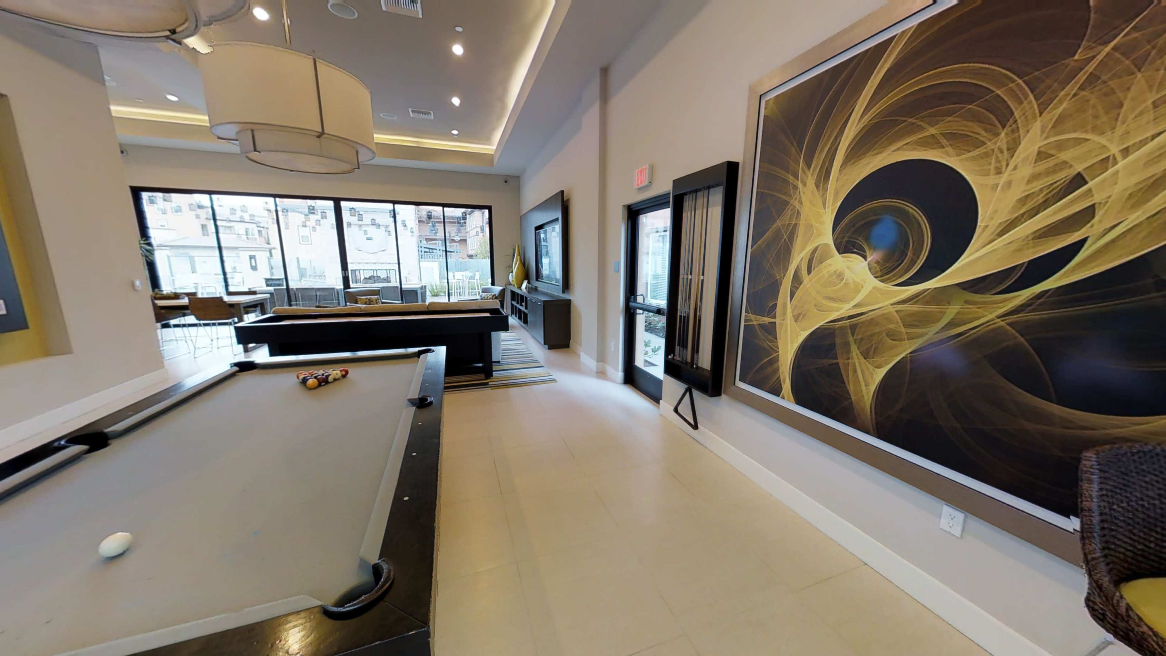 The image shows a modern lounge area featuring a pool table, a large abstract painting, and floor-to-ceiling windows.