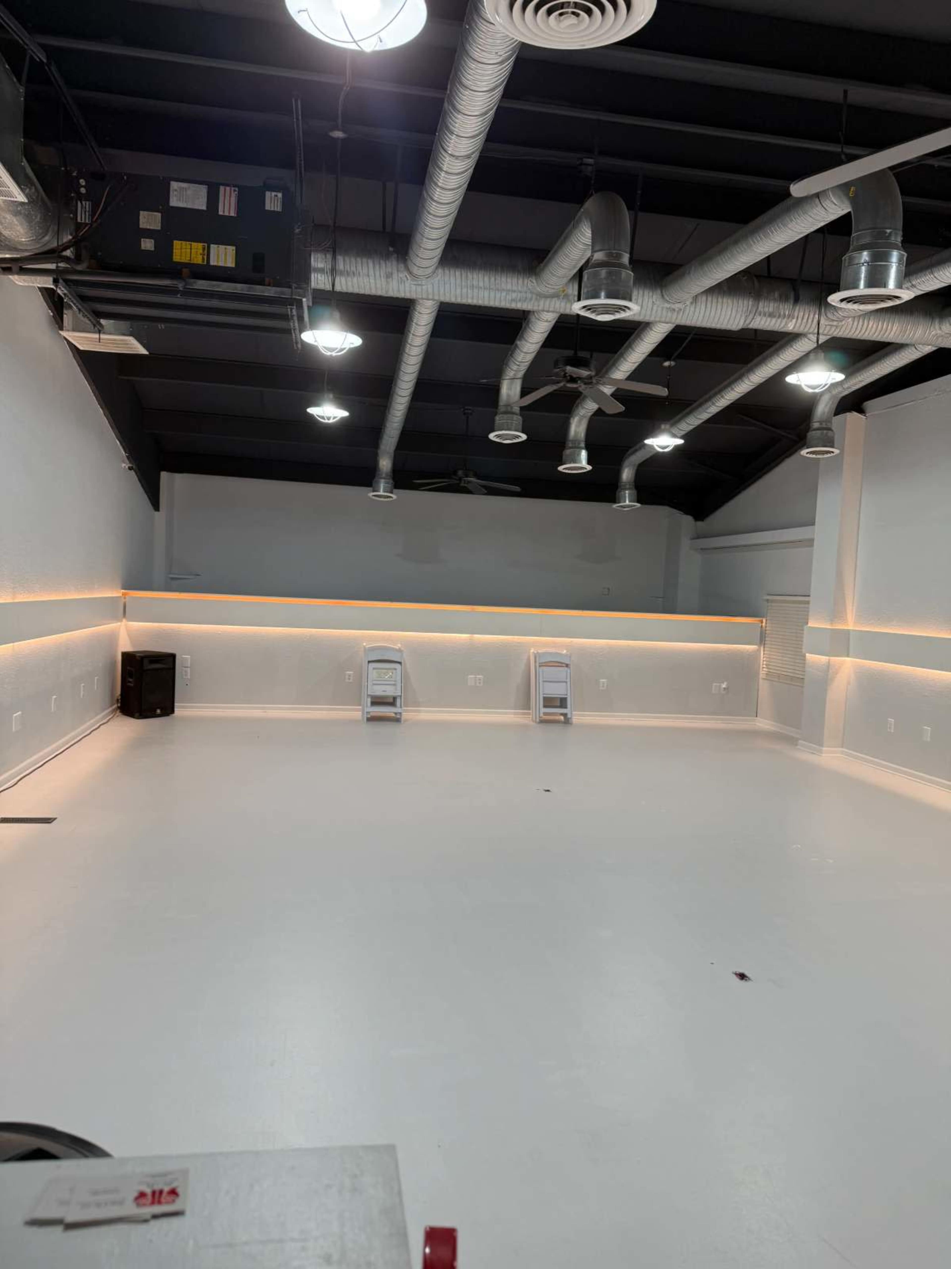 The image shows a spacious, empty room with white walls and a white floor, featuring overhead lights and exposed ductwork.