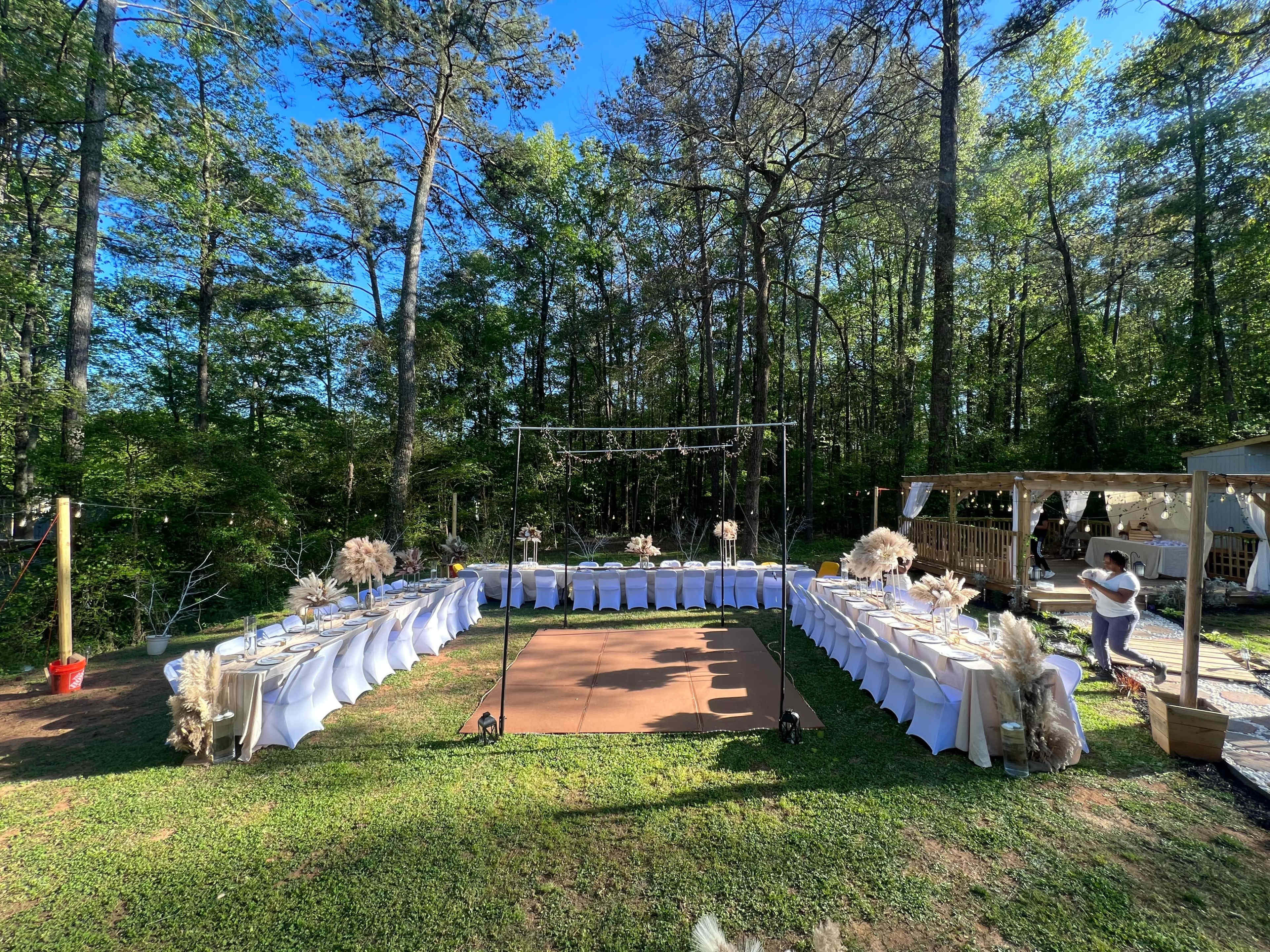 A rectangular outdoor event setup features tables arranged in a curve, surrounded by trees, with a dance floor and a decorated arch in the center.