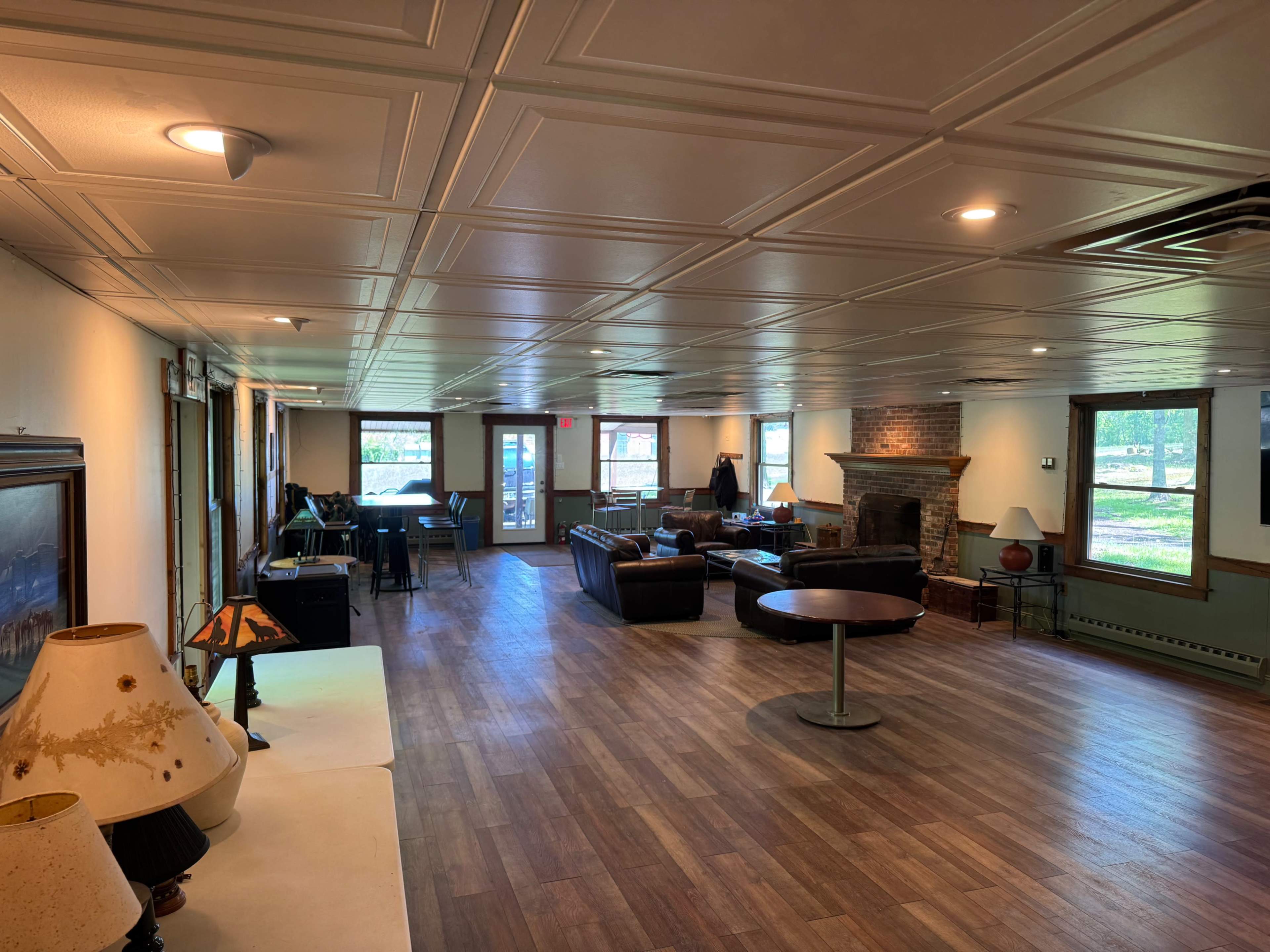 The image shows a spacious room with wooden flooring, featuring couches, a coffee table, a fireplace, and a dining area near a set of large windows.