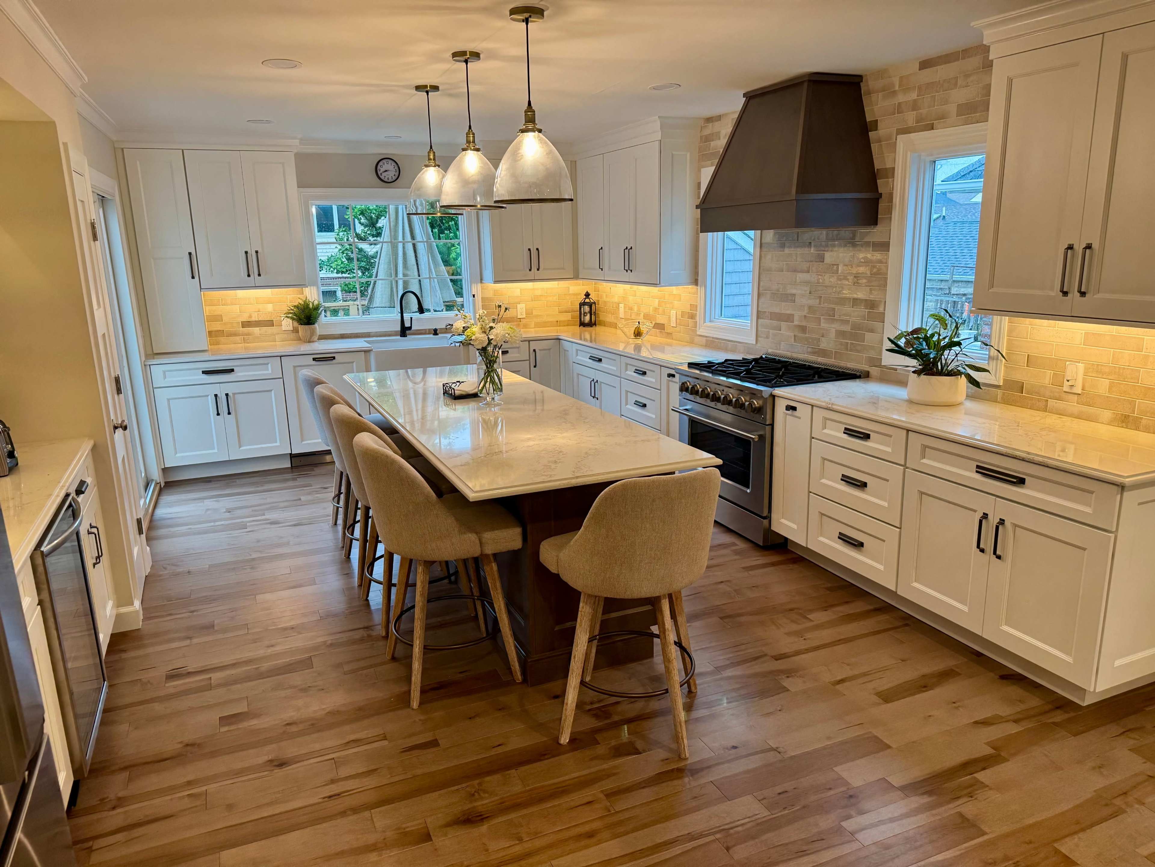 The image shows a modern kitchen with a large island, six barstools, white cabinets, and a gas range under a hood, illuminated by pendant lights.