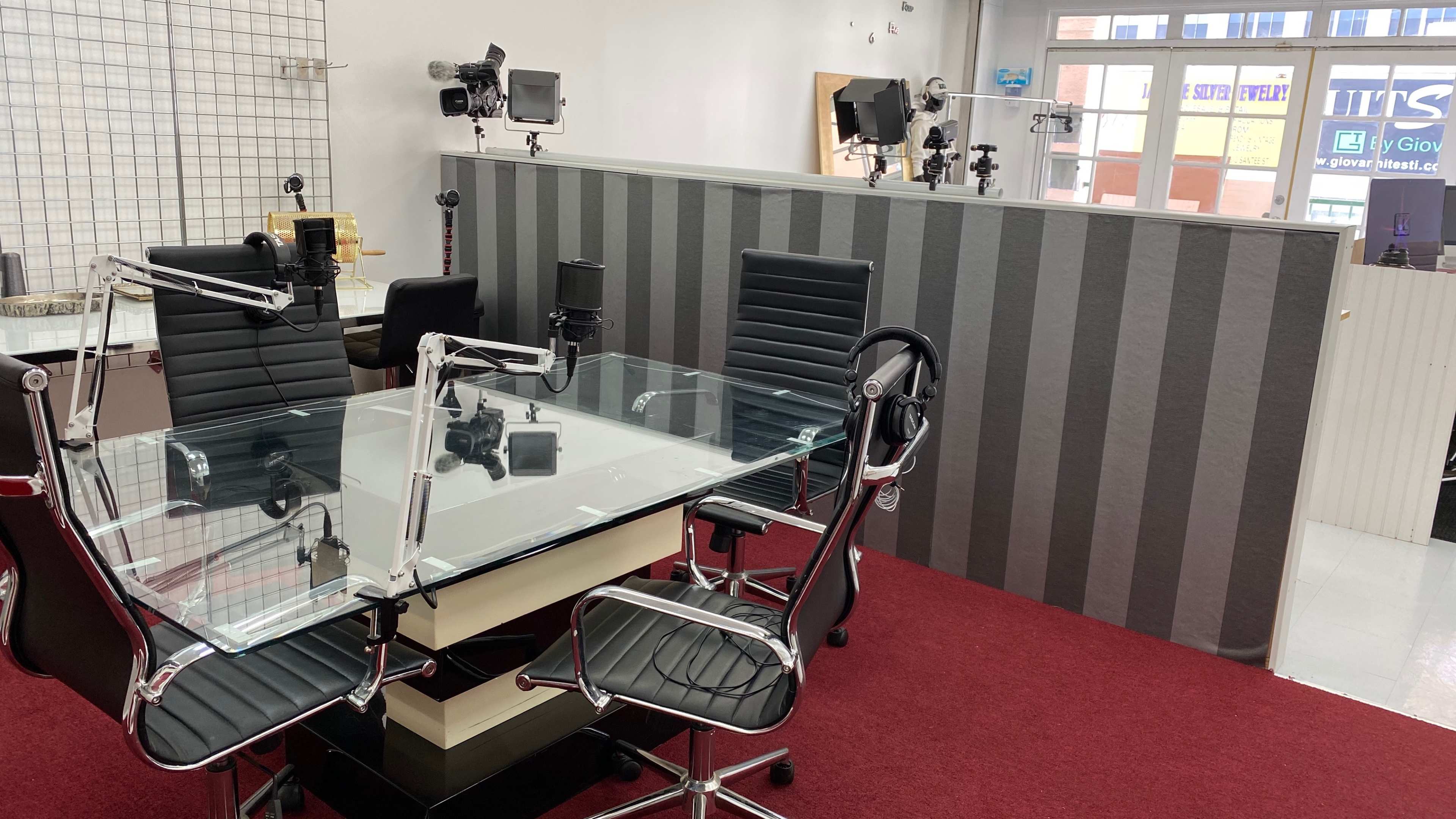 A modern recording studio with a glass table surrounded by black chairs and microphone setups, situated against a wall with grey stripes.